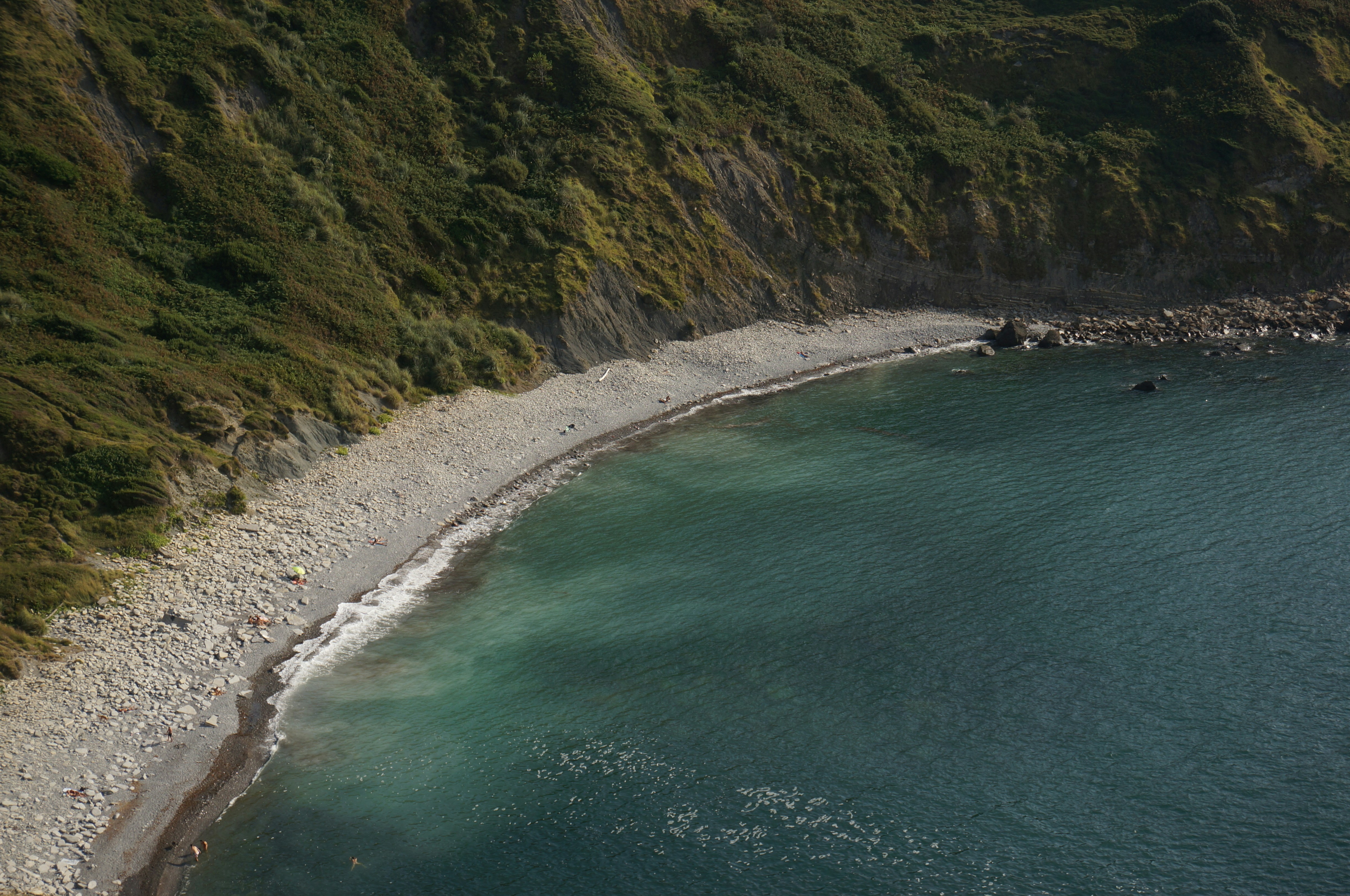 A pebble beach below a steep green cliff meeting the deep blue sea