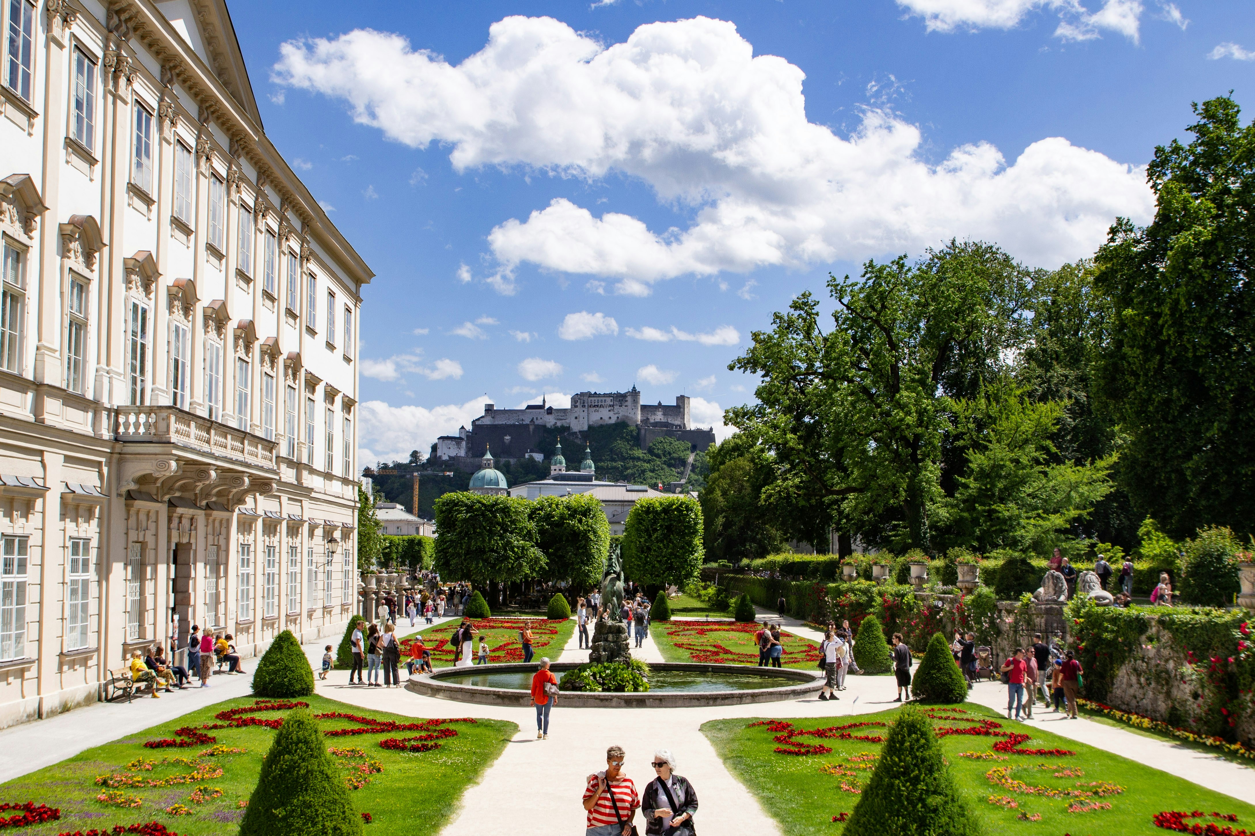 Orang-orang berjalan melalui Taman Mirabell dengan Benteng Hohensalzburg di latar belakang Salzburg