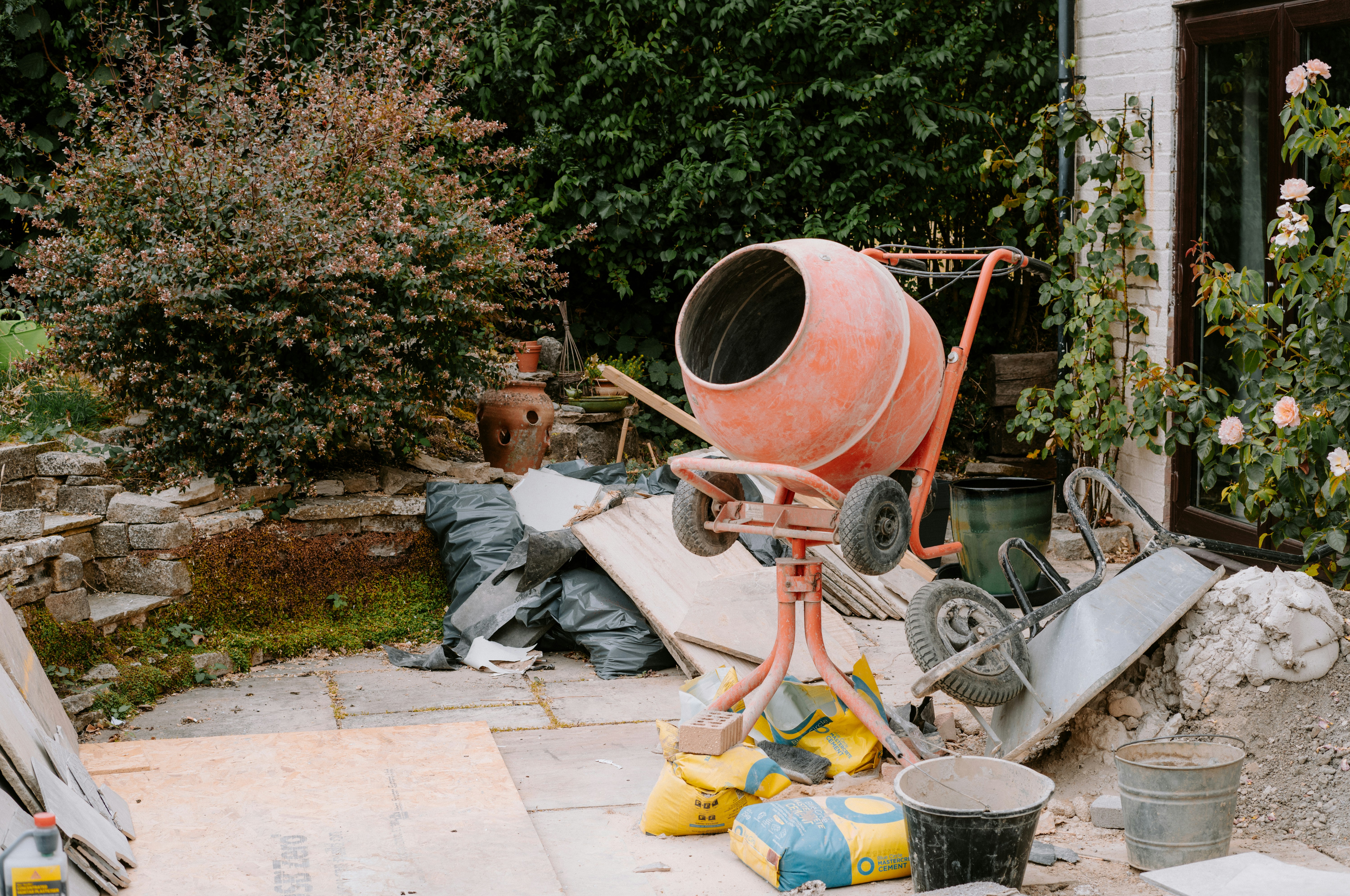 Ein orangefarbener Zementmischer auf einer Terrasse mit Baumaterialien und Gartenpflanzen