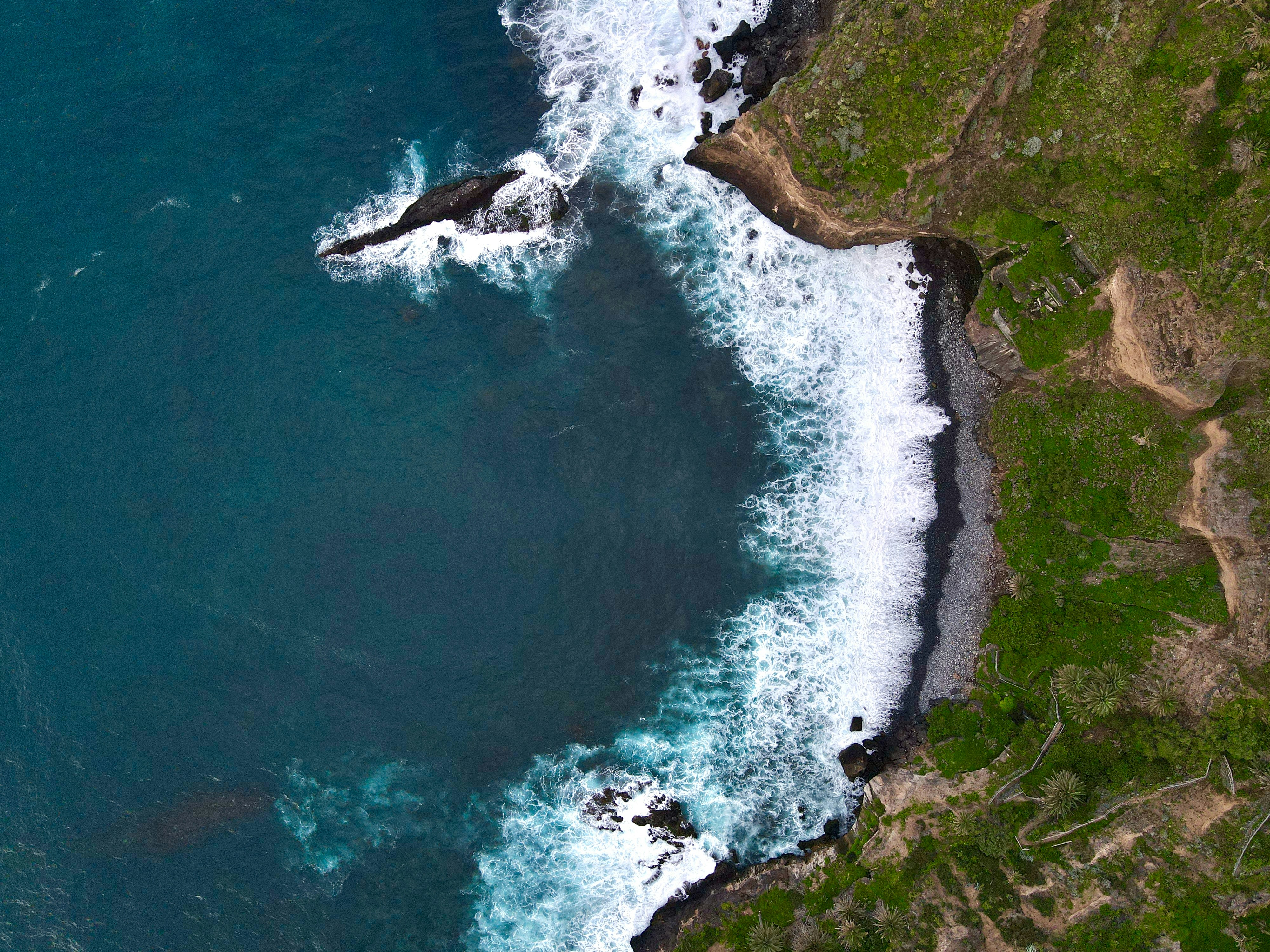 An aerial view of white waves crashing against a black sand beach and cliffs