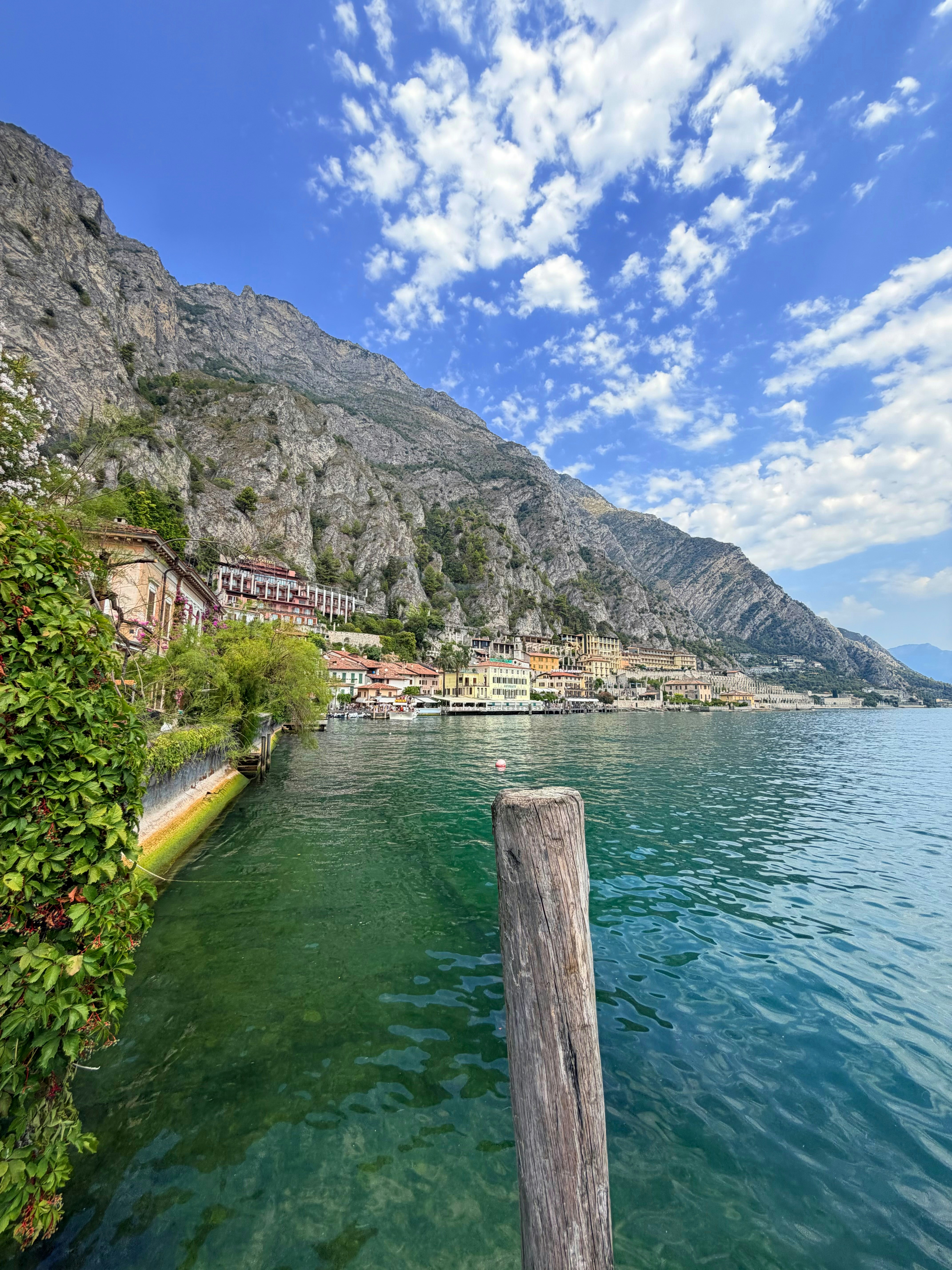 A wooden post in the green water of Lake Garda, Italy, with mountains behind