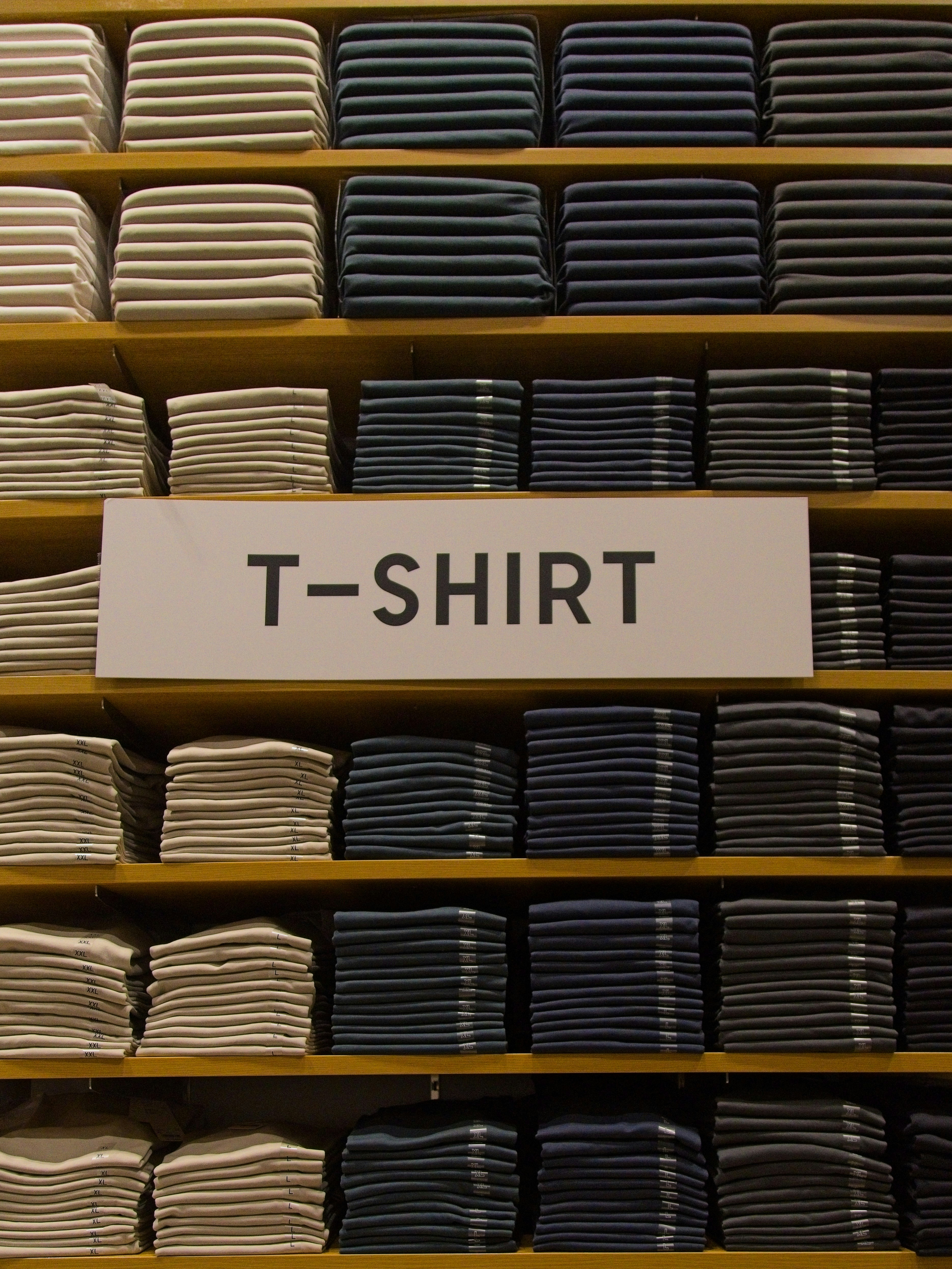 Stacks of folded t-shirts in neutral colors on wooden shelves with a sign