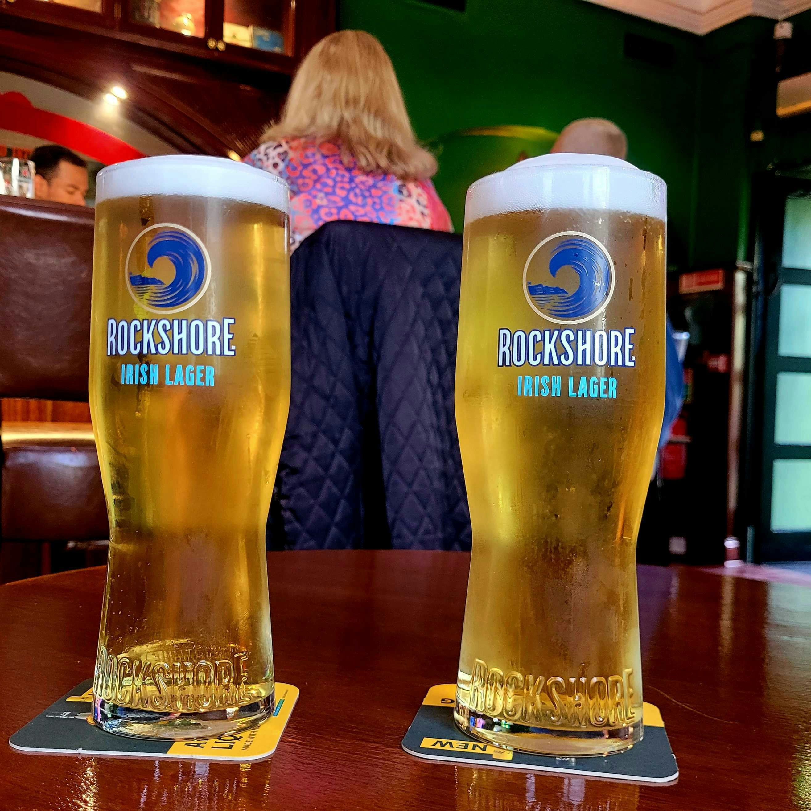 Dua gelas Rockshore Irish Lager di atas tatakan gelas di meja pub kayu
