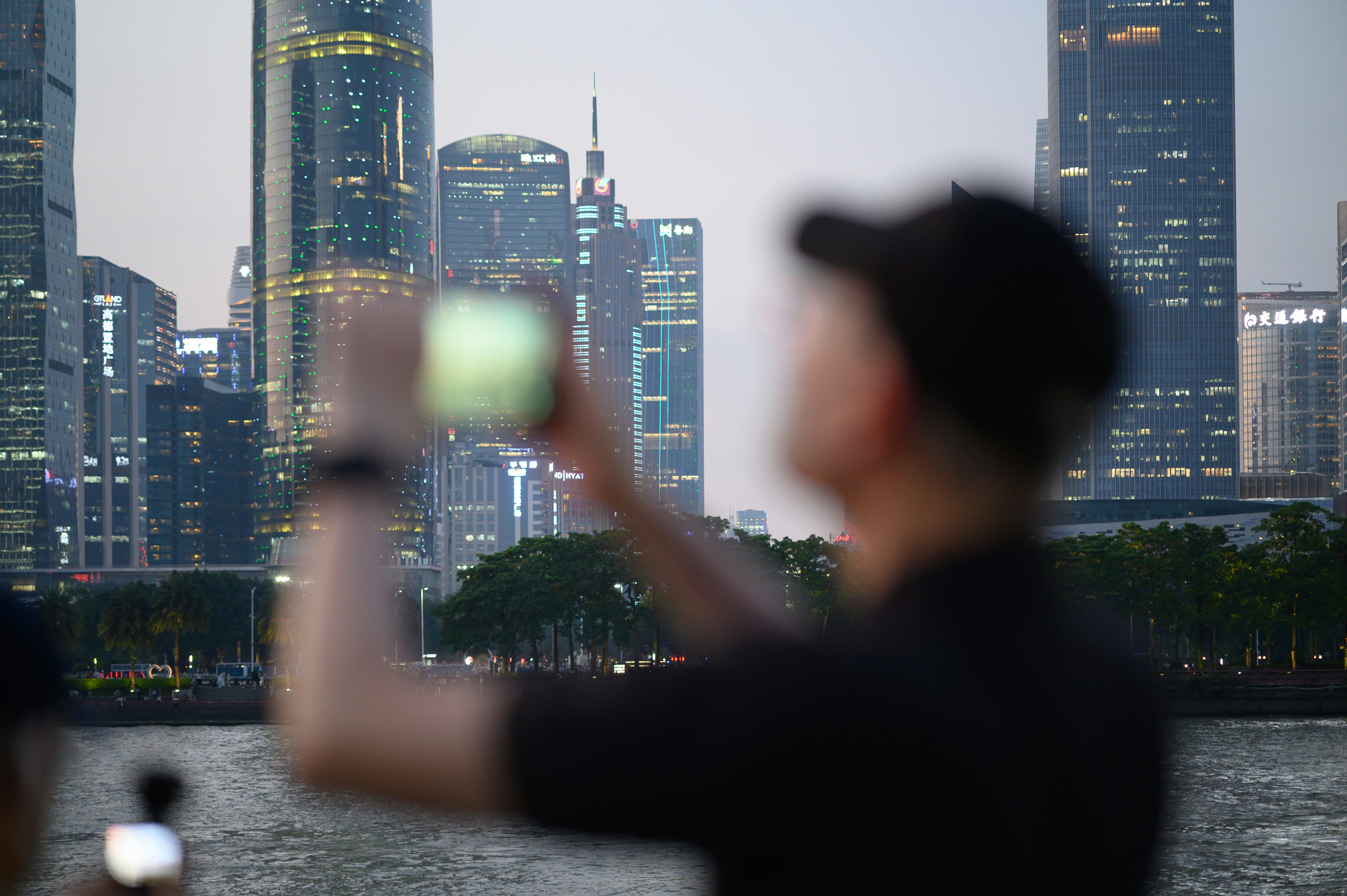 A person in a black shirt photographing a city skyline across a river.