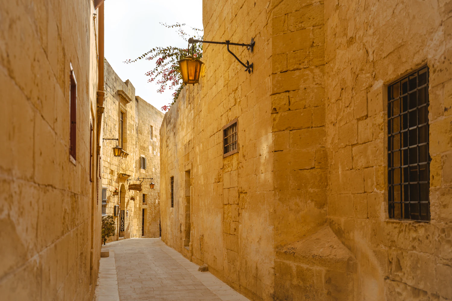 A narrow limestone lane in hard sunlight, a wrought-iron lantern on the wall and shuttered windows beyond