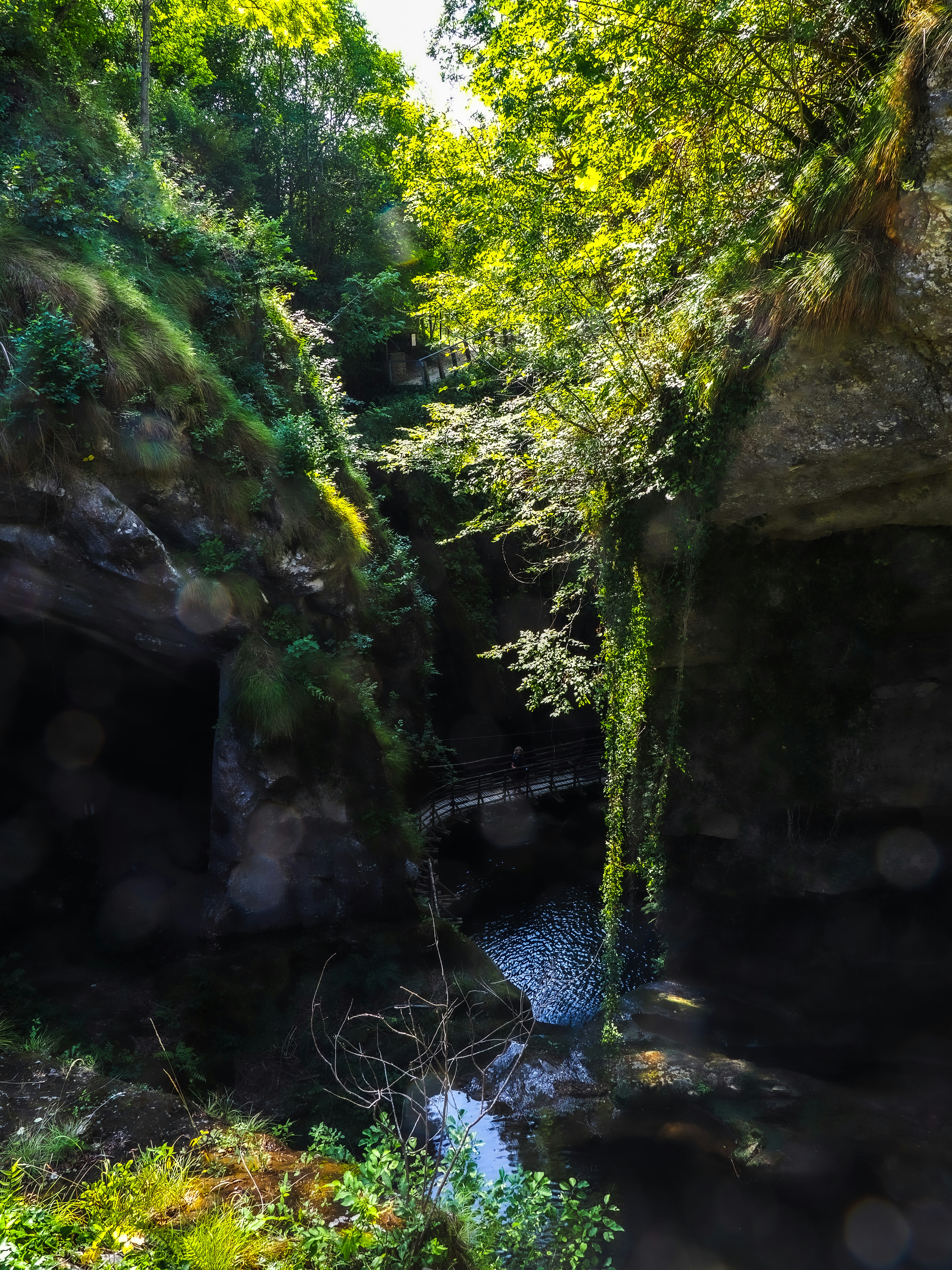 A narrow wooden footbridge crossing a dark pool between steep, mossy rock walls