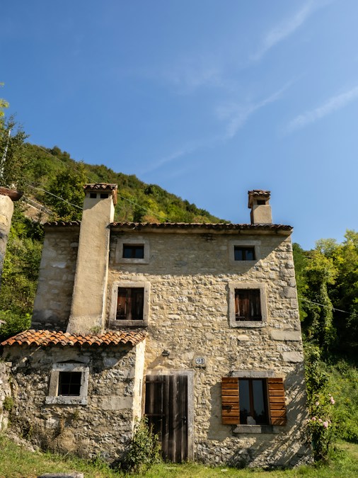Maison de campagne en pierre aux volets de bois, au pied d’une colline boisée sous un ciel bleu