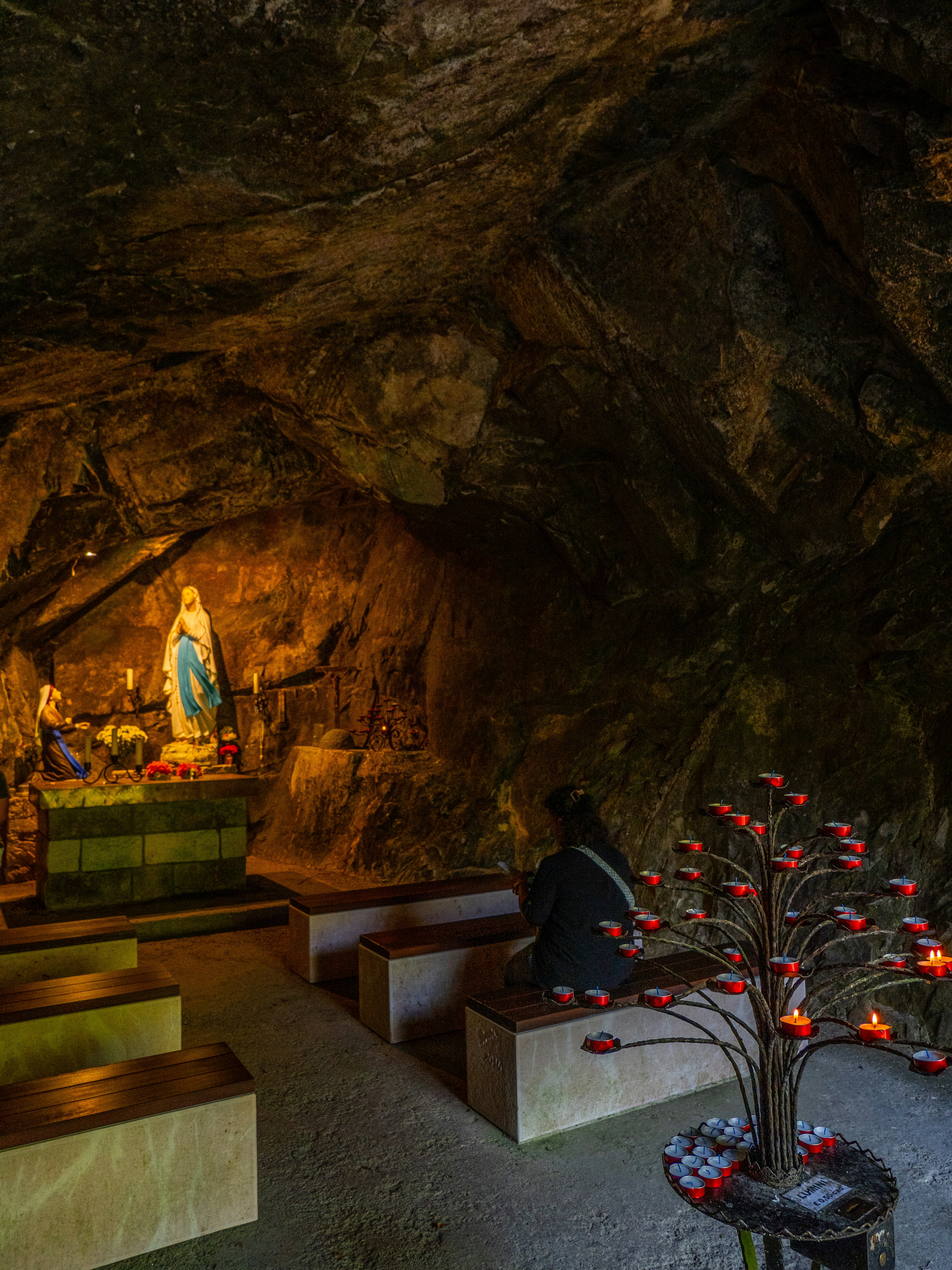 A person sitting on a bench in a cave shrine with a statue and candles
