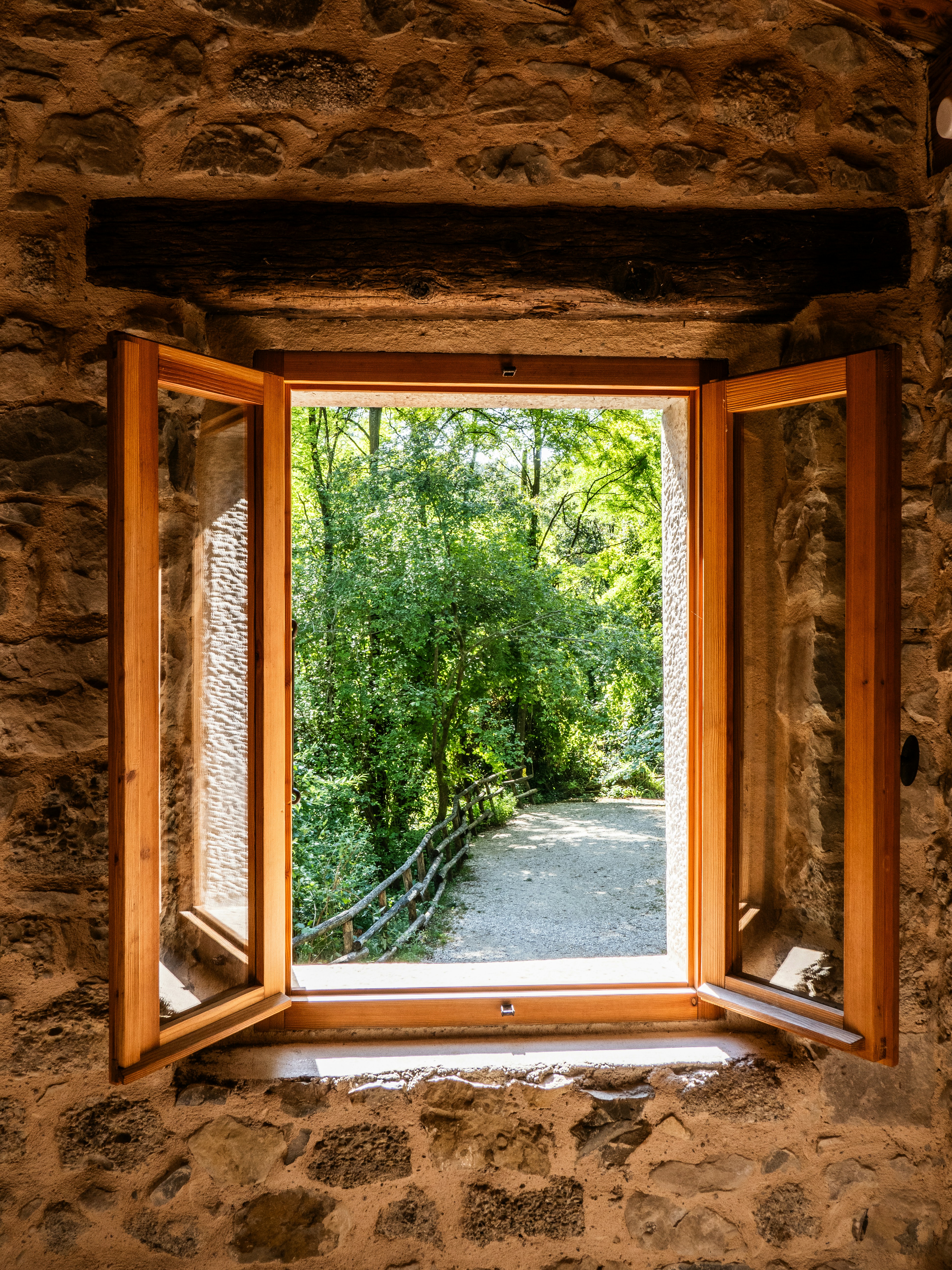 An open wooden window looking out onto a gravel path and green trees