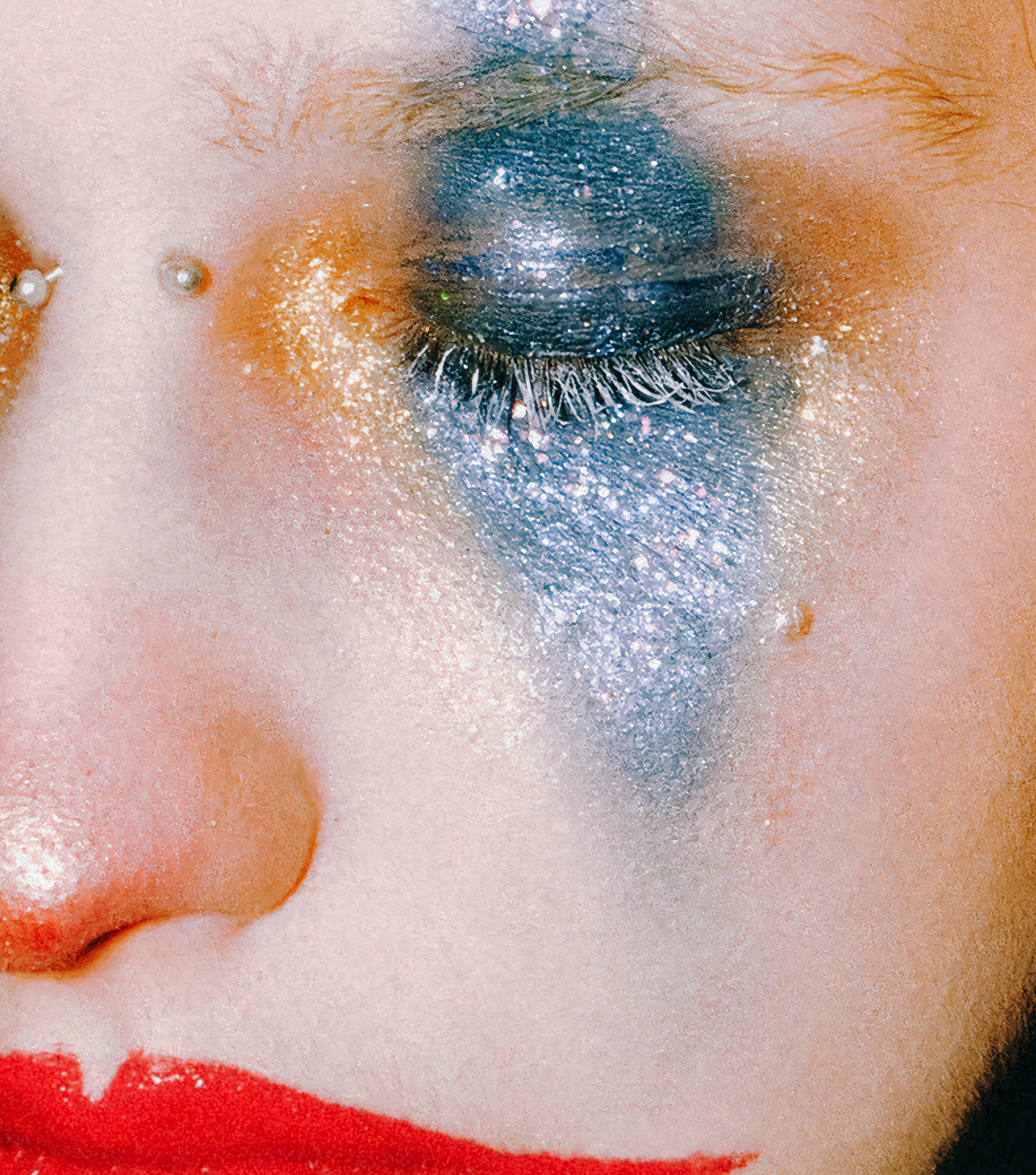 A person with blue glitter eyeshadow, gold accents, and bright red lipstick.