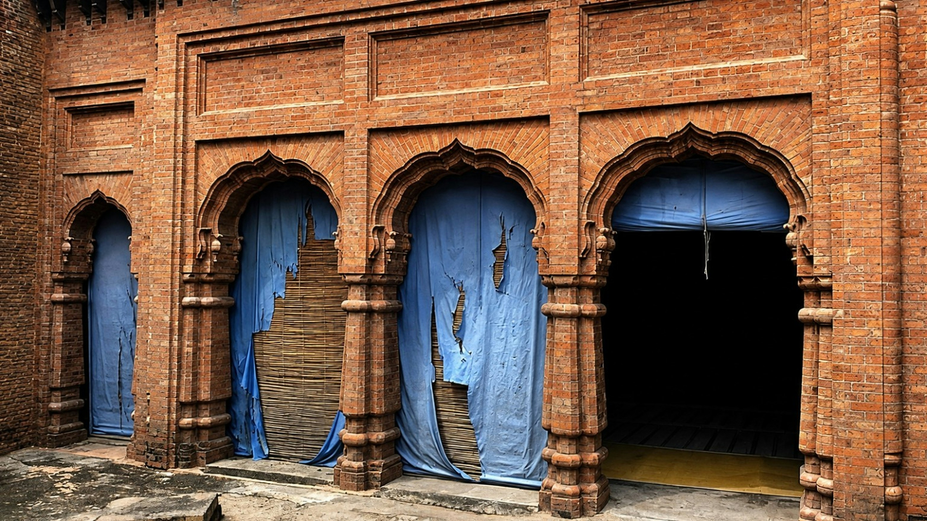 Three arched brick doorways with torn blue fabric coverings and bamboo screens.