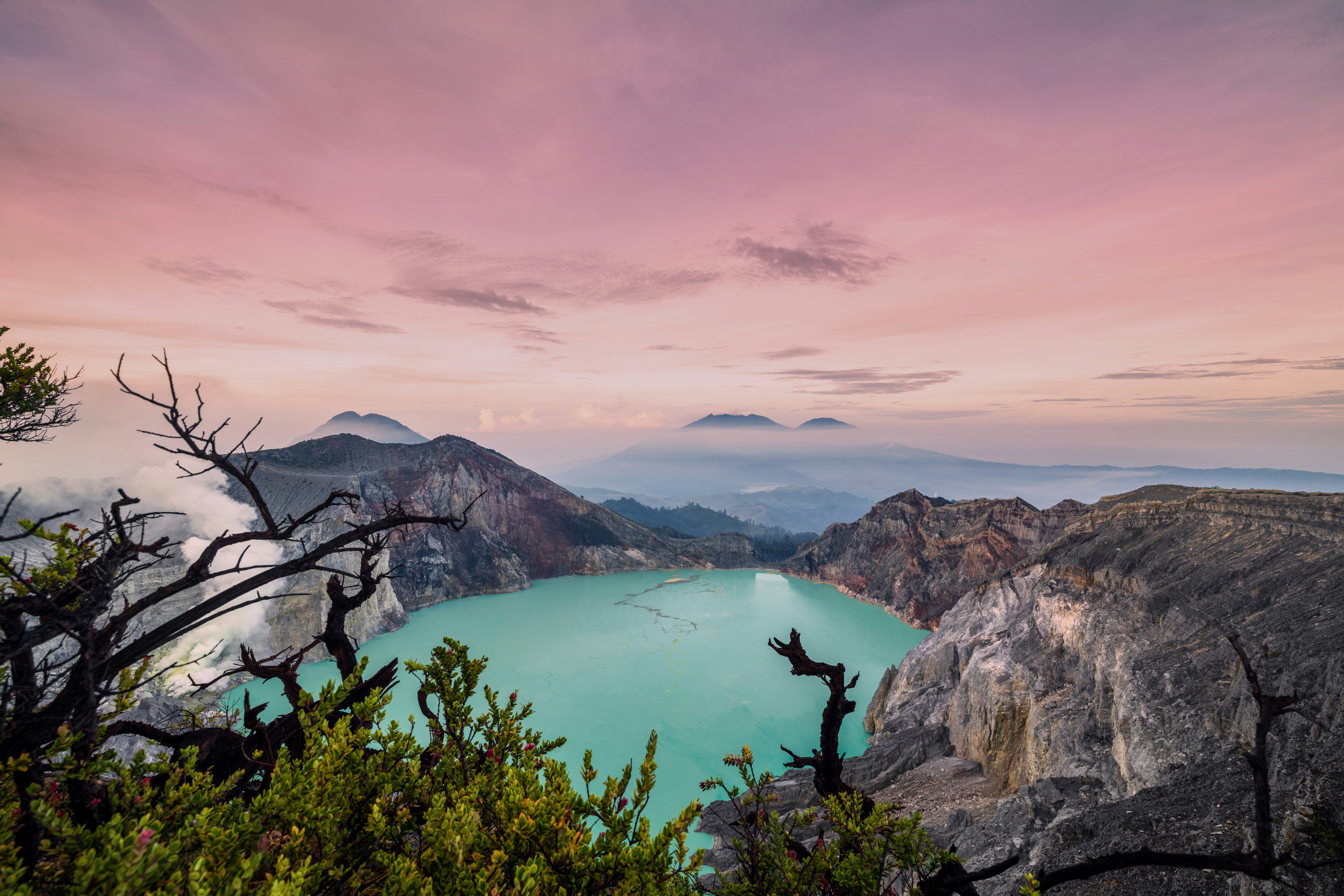 A turquoise volcanic crater lake surrounded by rocky cliffs under a soft pink sky