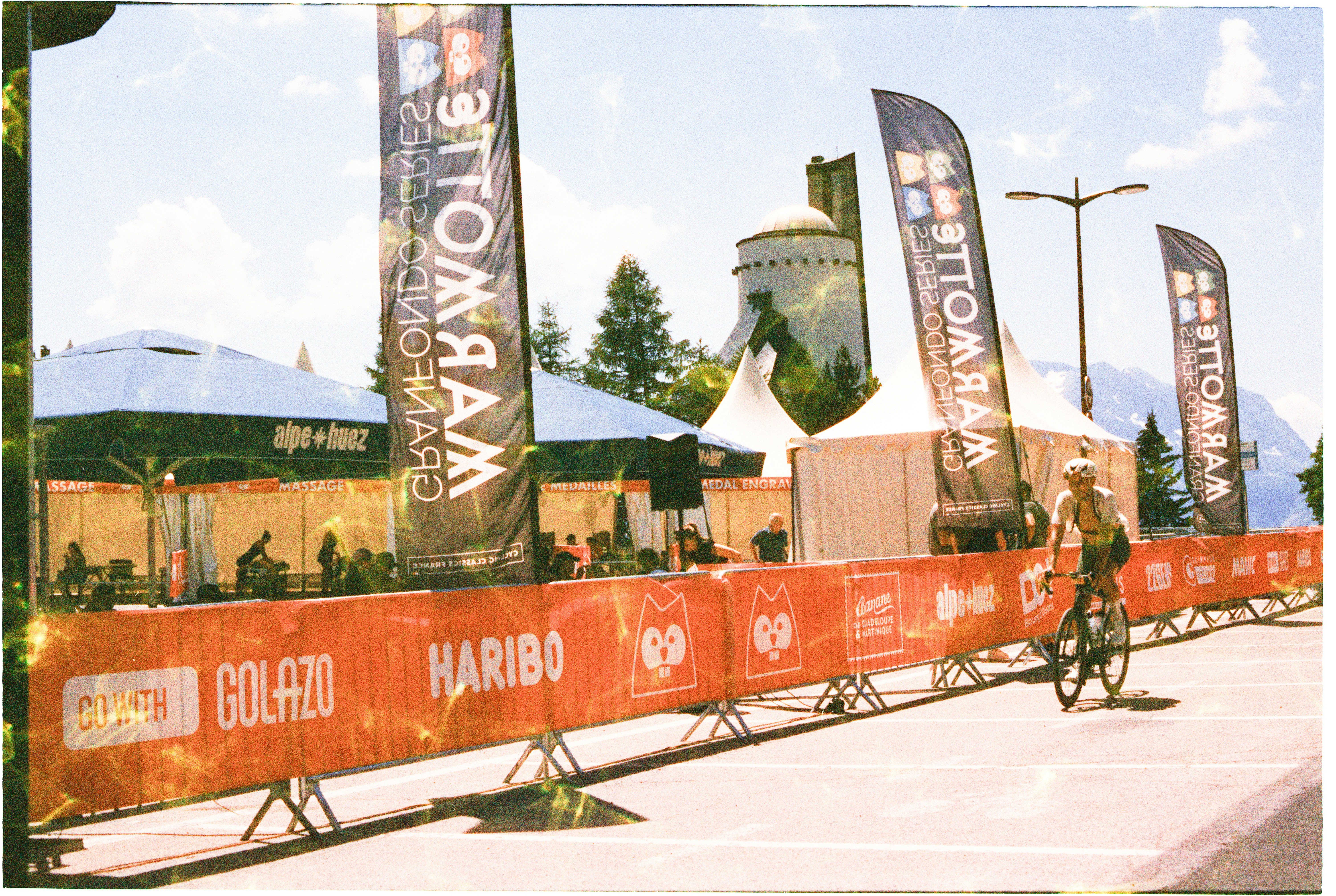 A cyclist riding past orange event barriers and tall banners in Alpe d'Huez