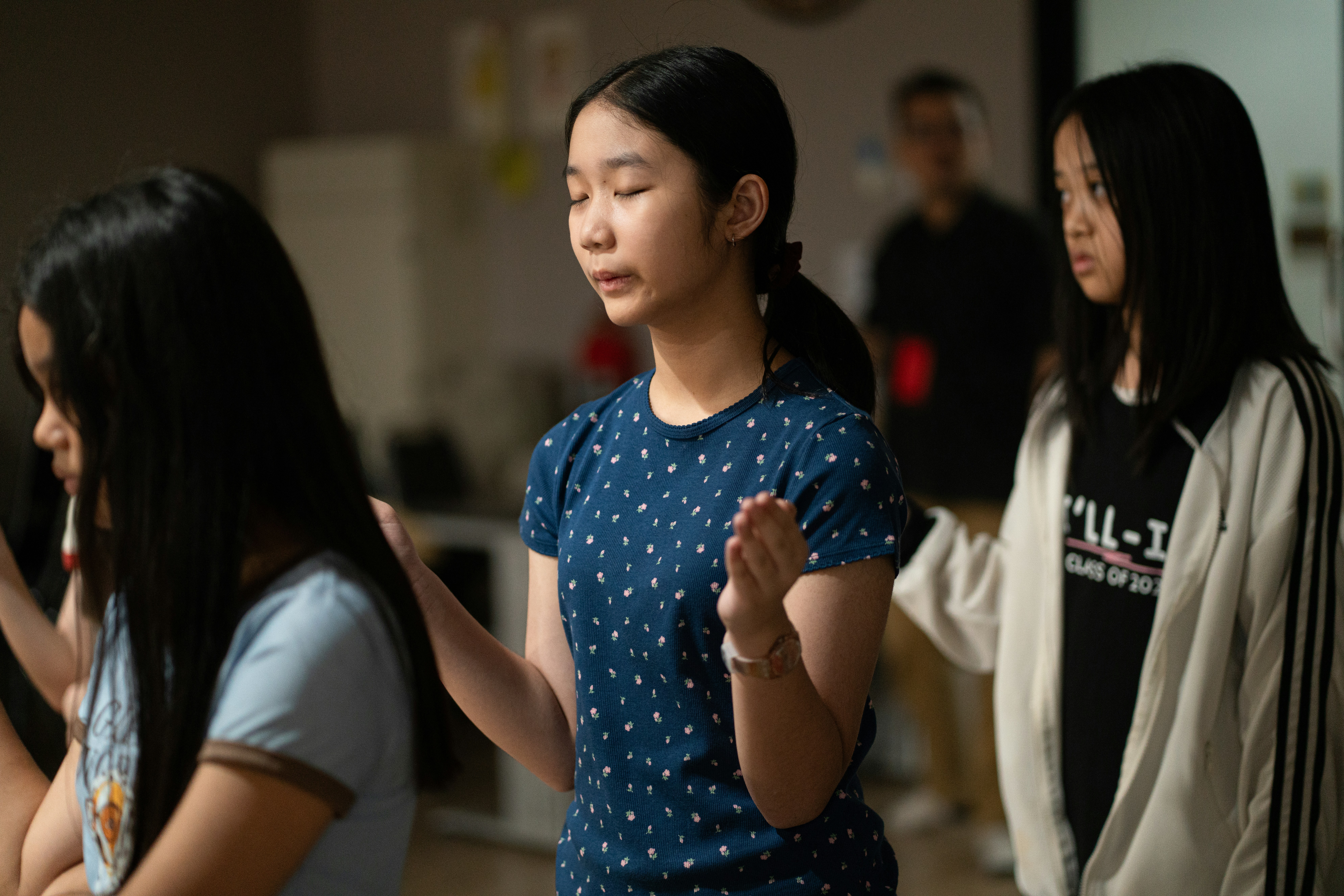 Three young people with eyes closed and hands raised in a quiet room.