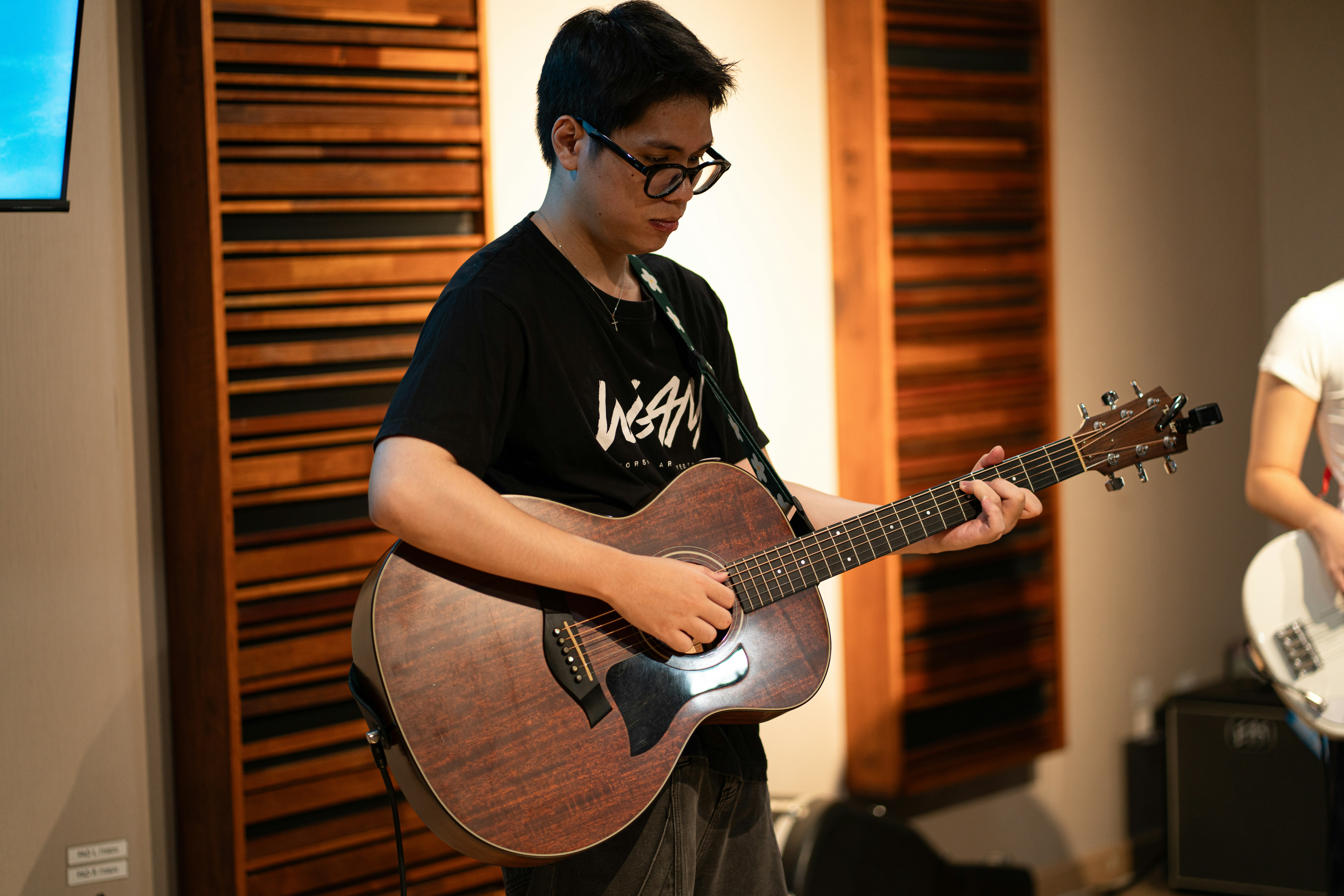 A person in glasses playing an acoustic guitar in a room with wooden slats.