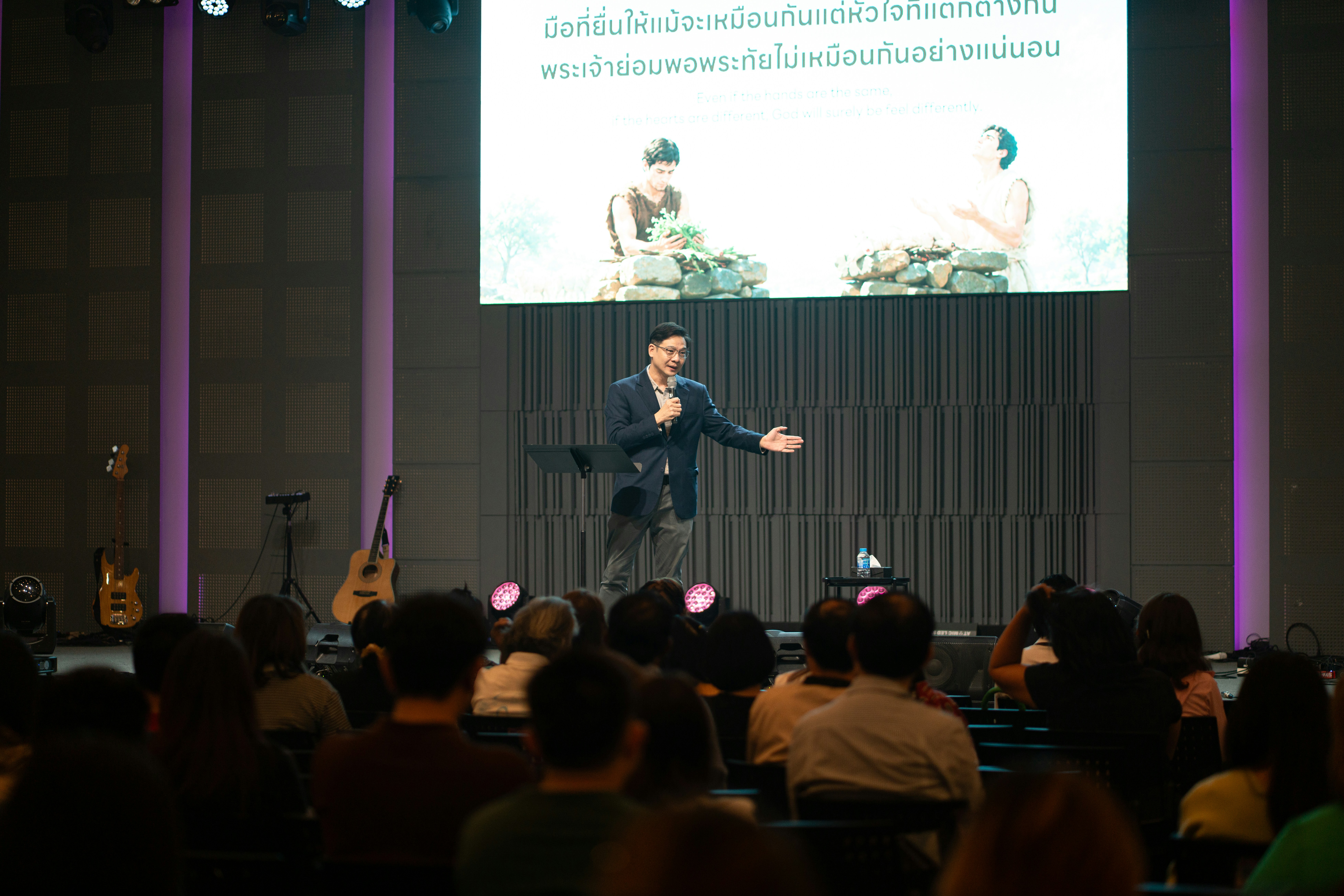 A man speaking into a microphone on stage before an audience in a hall.