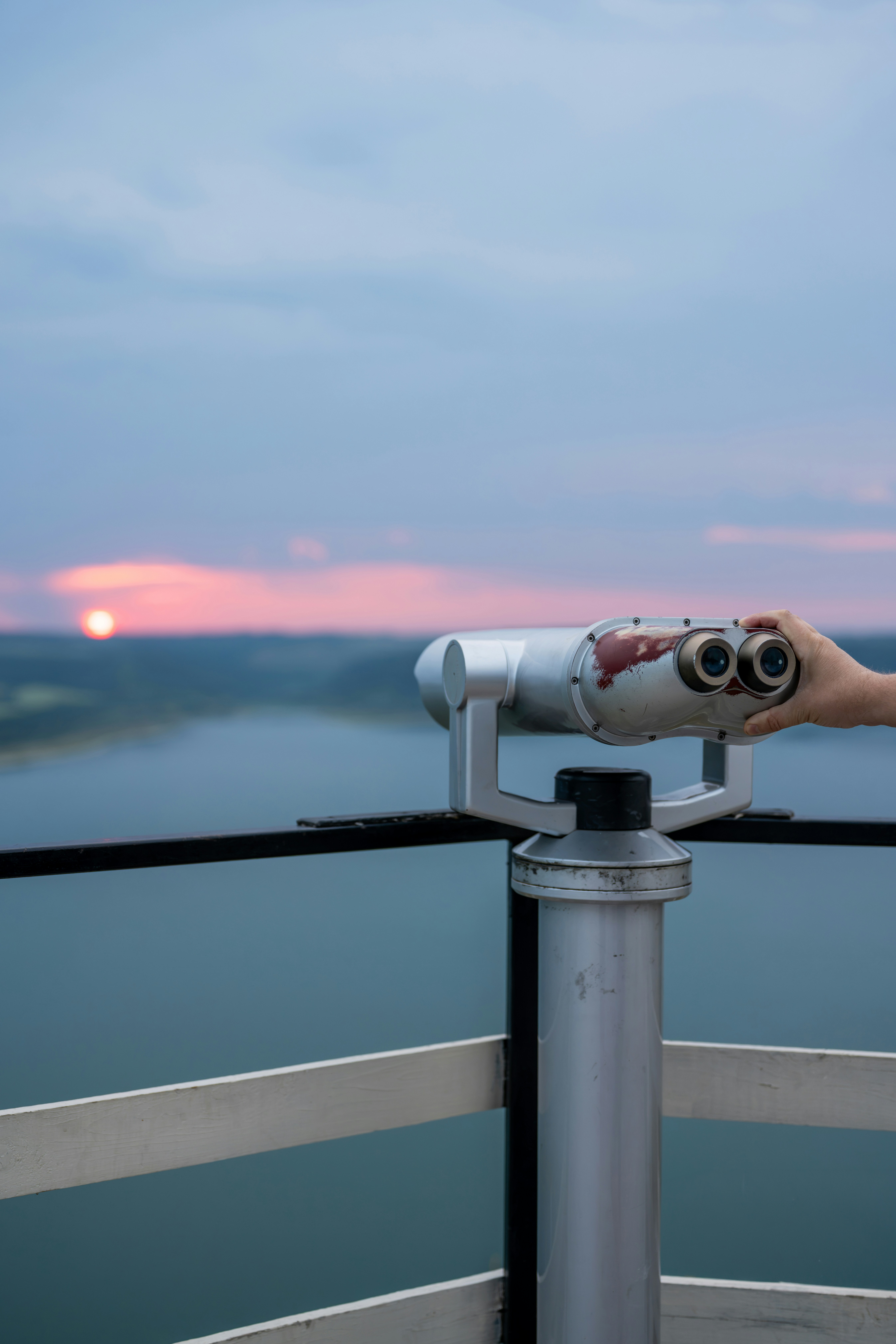 A hand adjusting a metal coin-operated binocular viewer overlooking a lake at low sun.