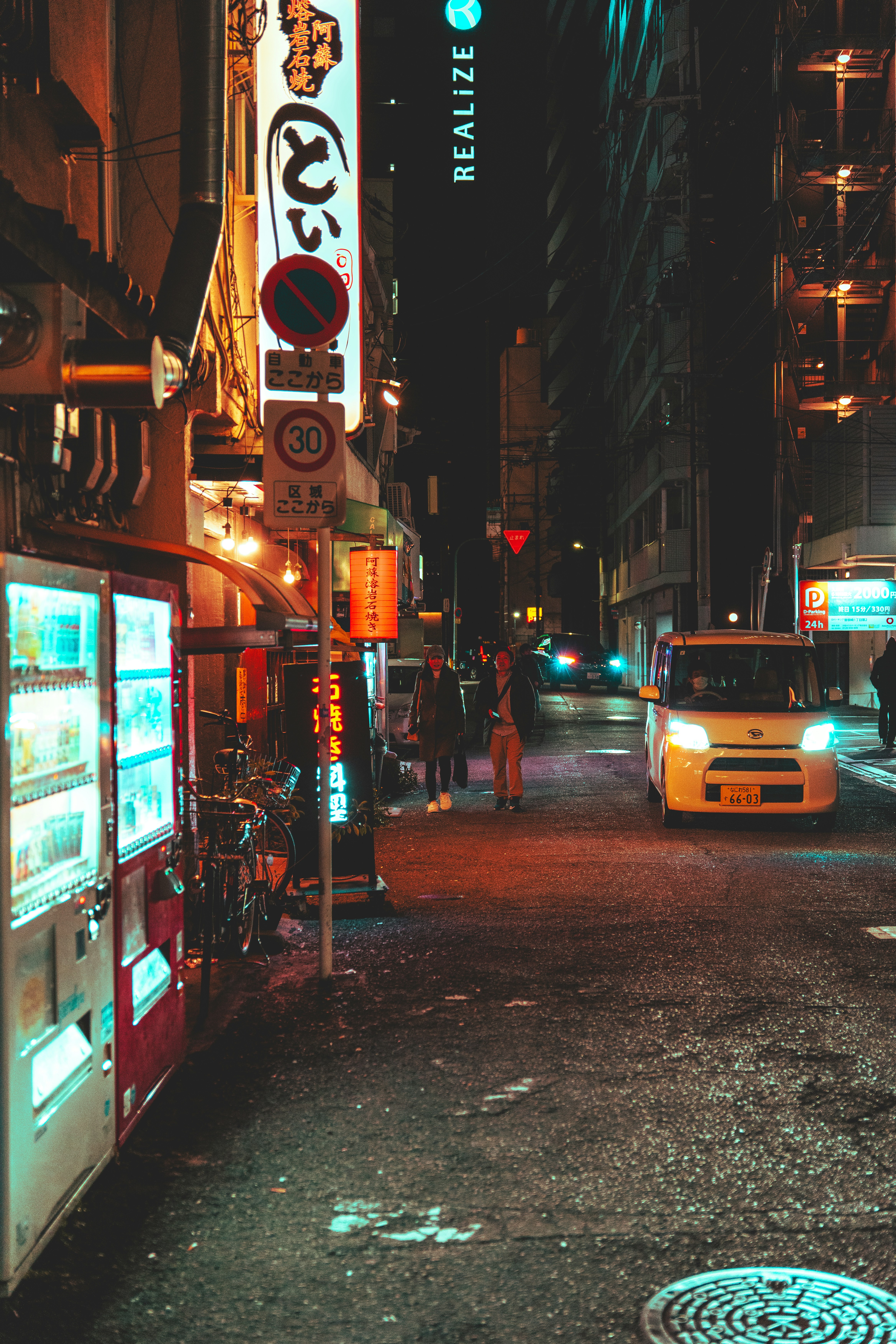 Two people walking down a narrow street at night with neon signs and a car