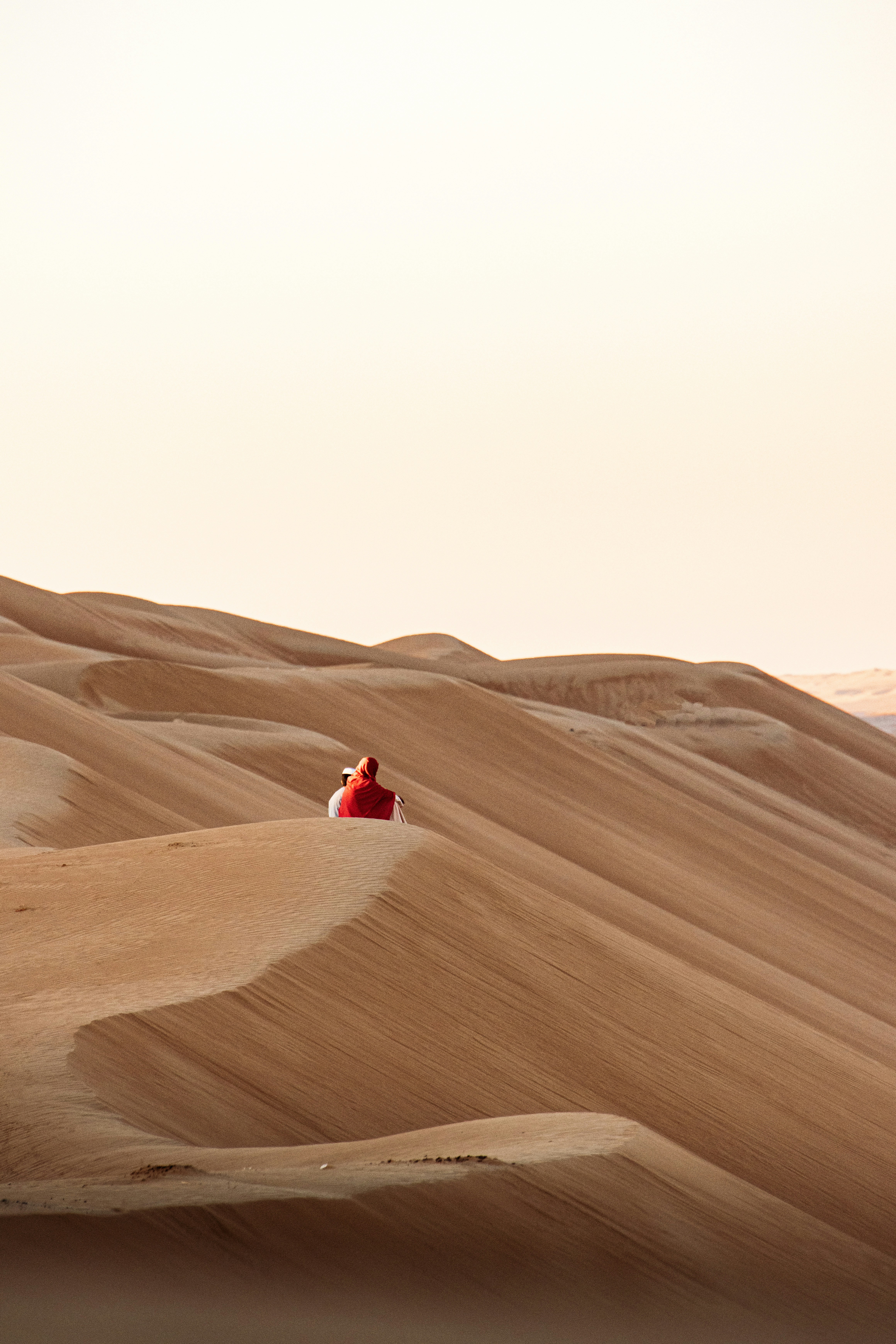 Two people in light clothing and a red shawl on a vast sand dune.