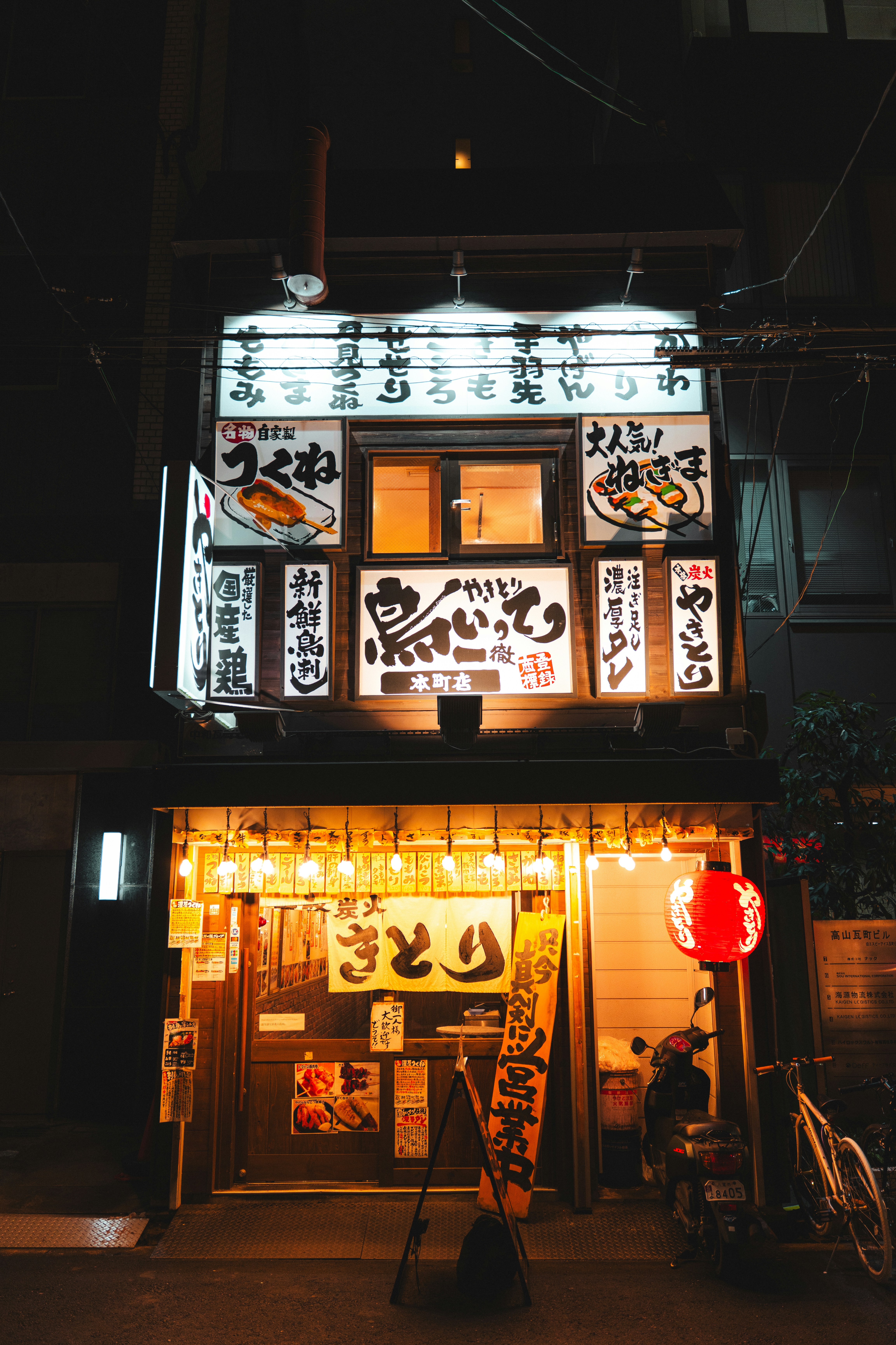 A yakitori restaurant storefront with illuminated signs and a red lantern at night.
