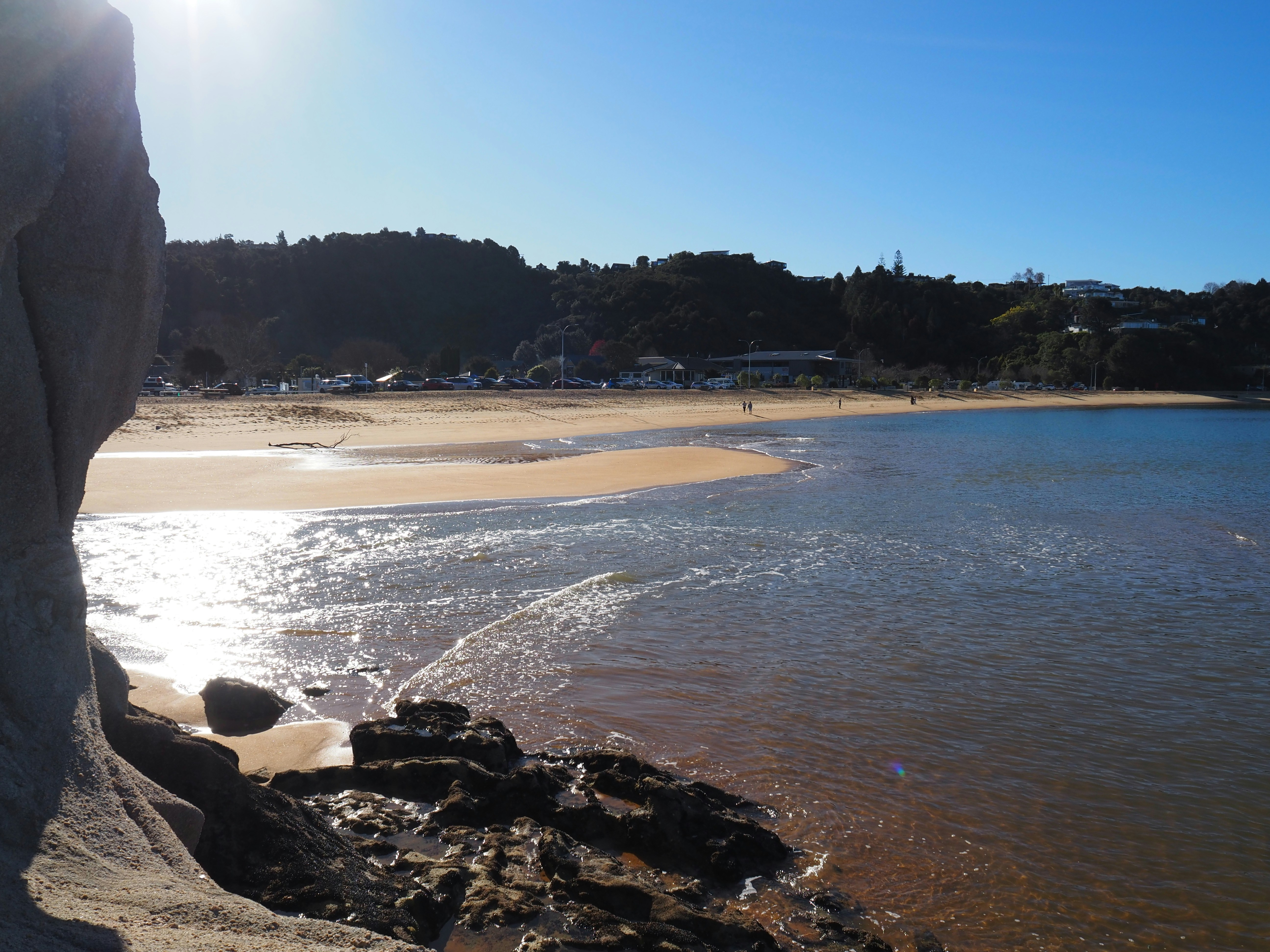 A sandy beach with dark rocks in the foreground under a clear blue sky
