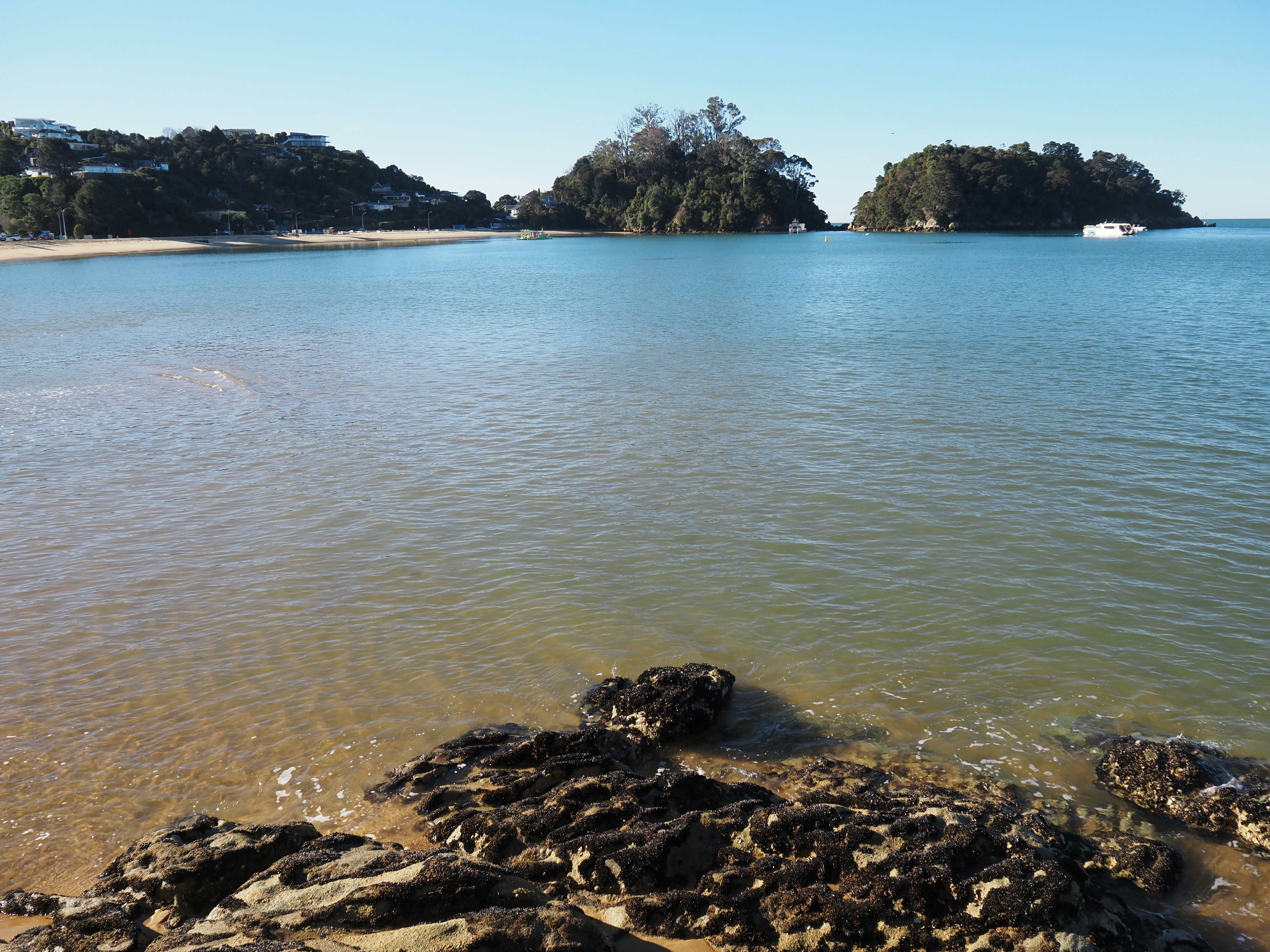 A calm bay with rocky shores, sandy beaches, and forested islands under clear skies