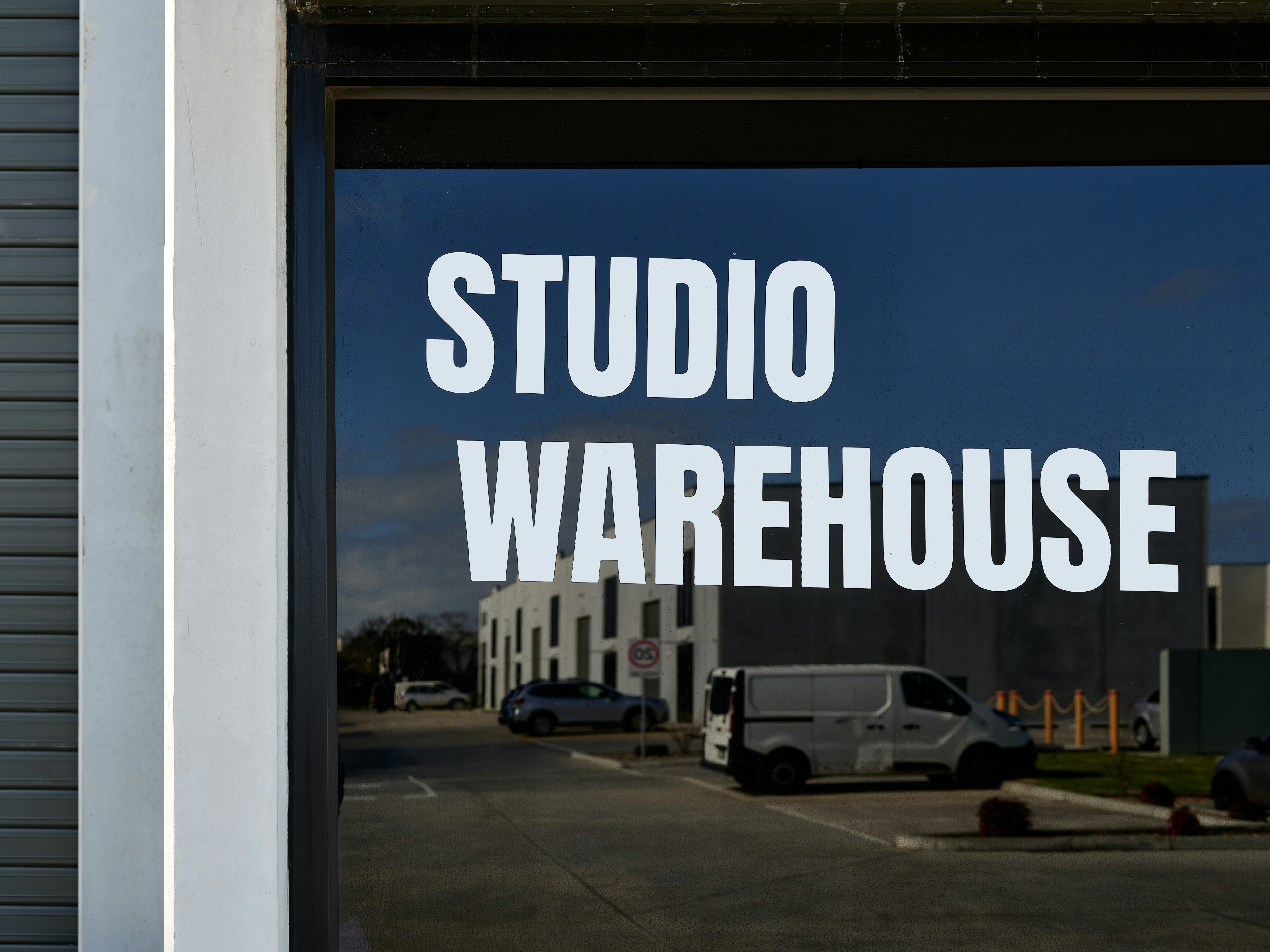 Pintu kaca dengan tulisan putih bertuliskan "STUDIO WAREHOUSE" yang menghadap ke tempat parkir