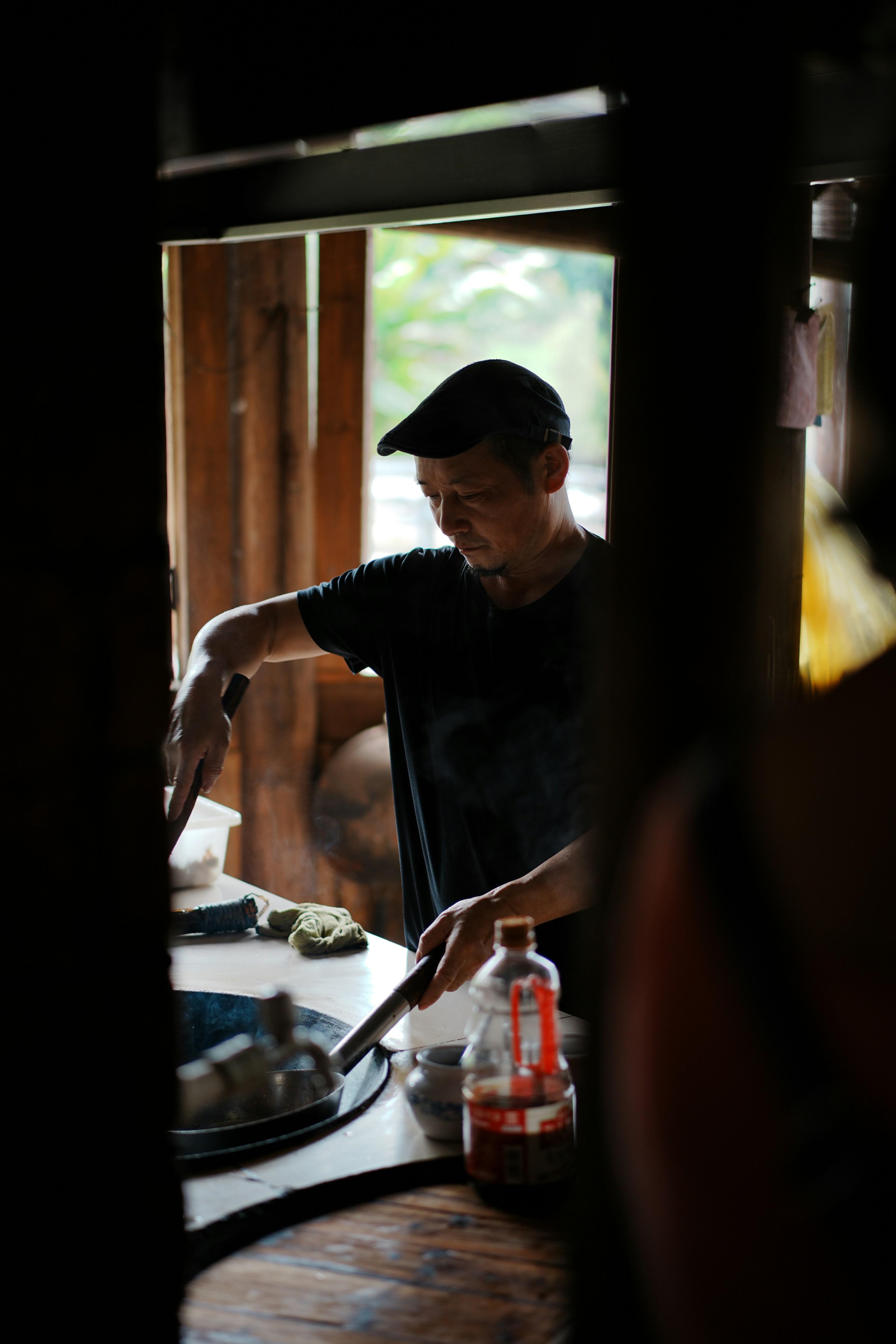 A man in a black t-shirt and cap cooking in a rustic kitchen.