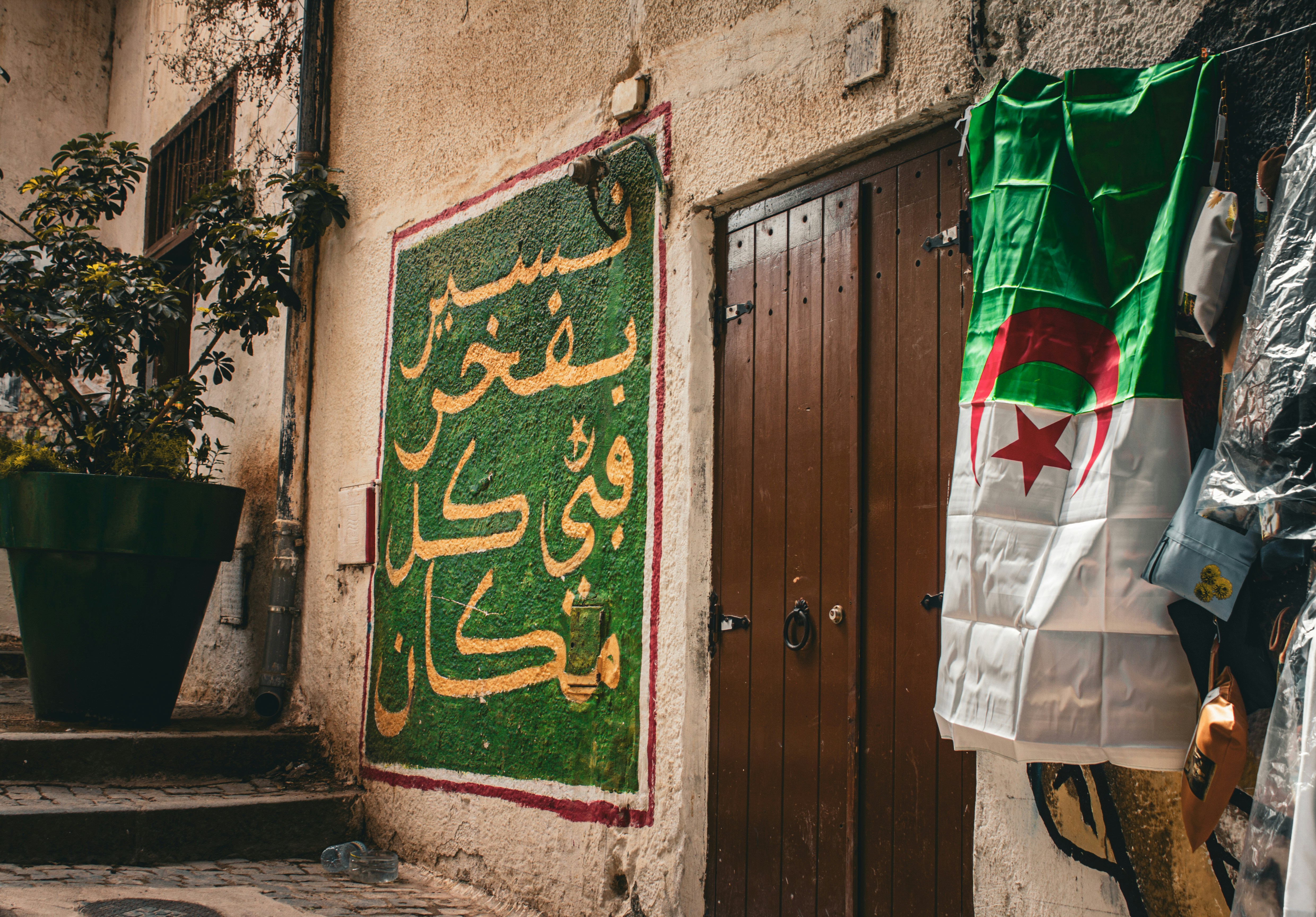 Drapeau algérien suspendu près d'une porte en bois et d'une fresque calligraphiée verte, dans une rue d'Alger