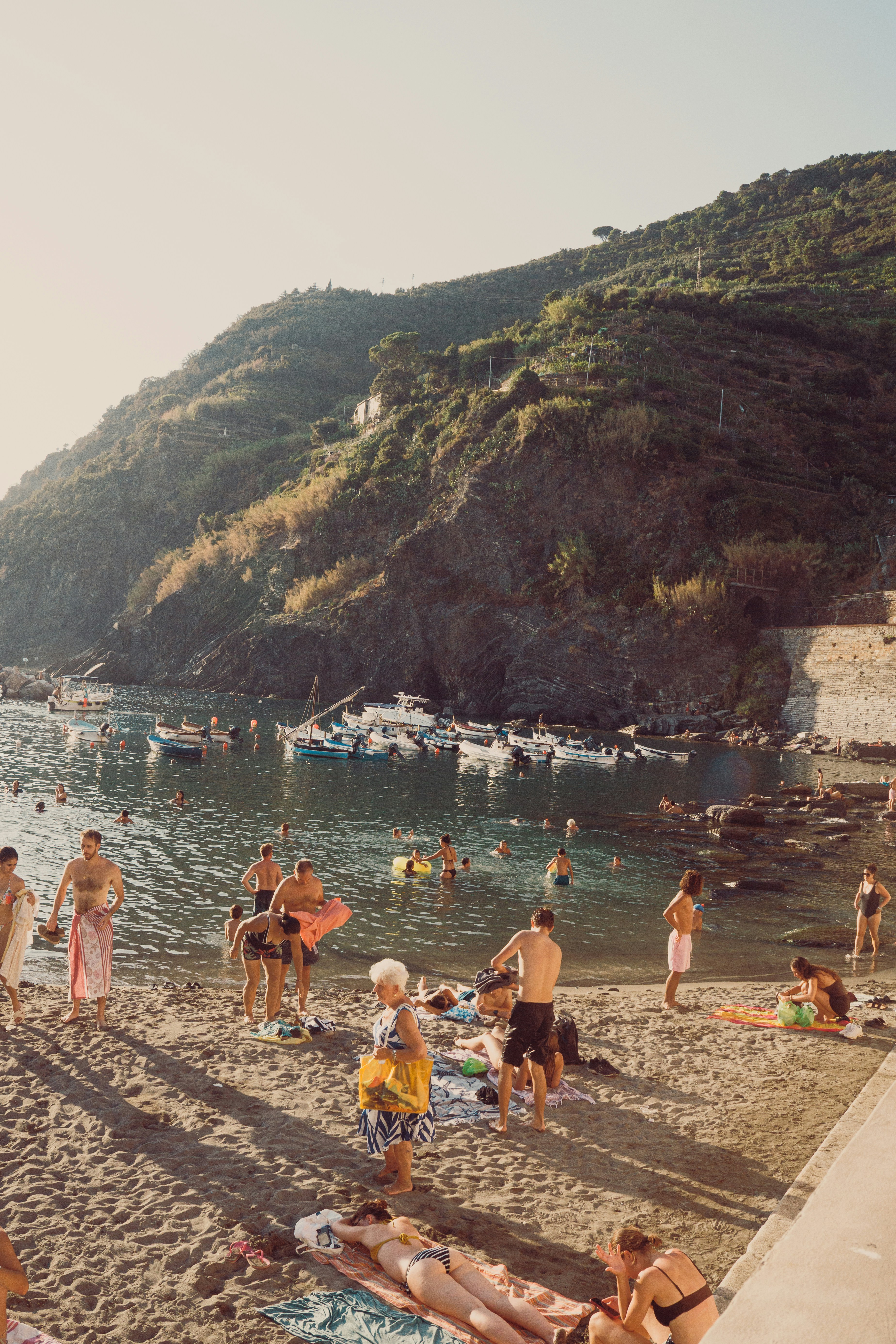 People on a sandy beach with boats in a bay below a rocky cliff.