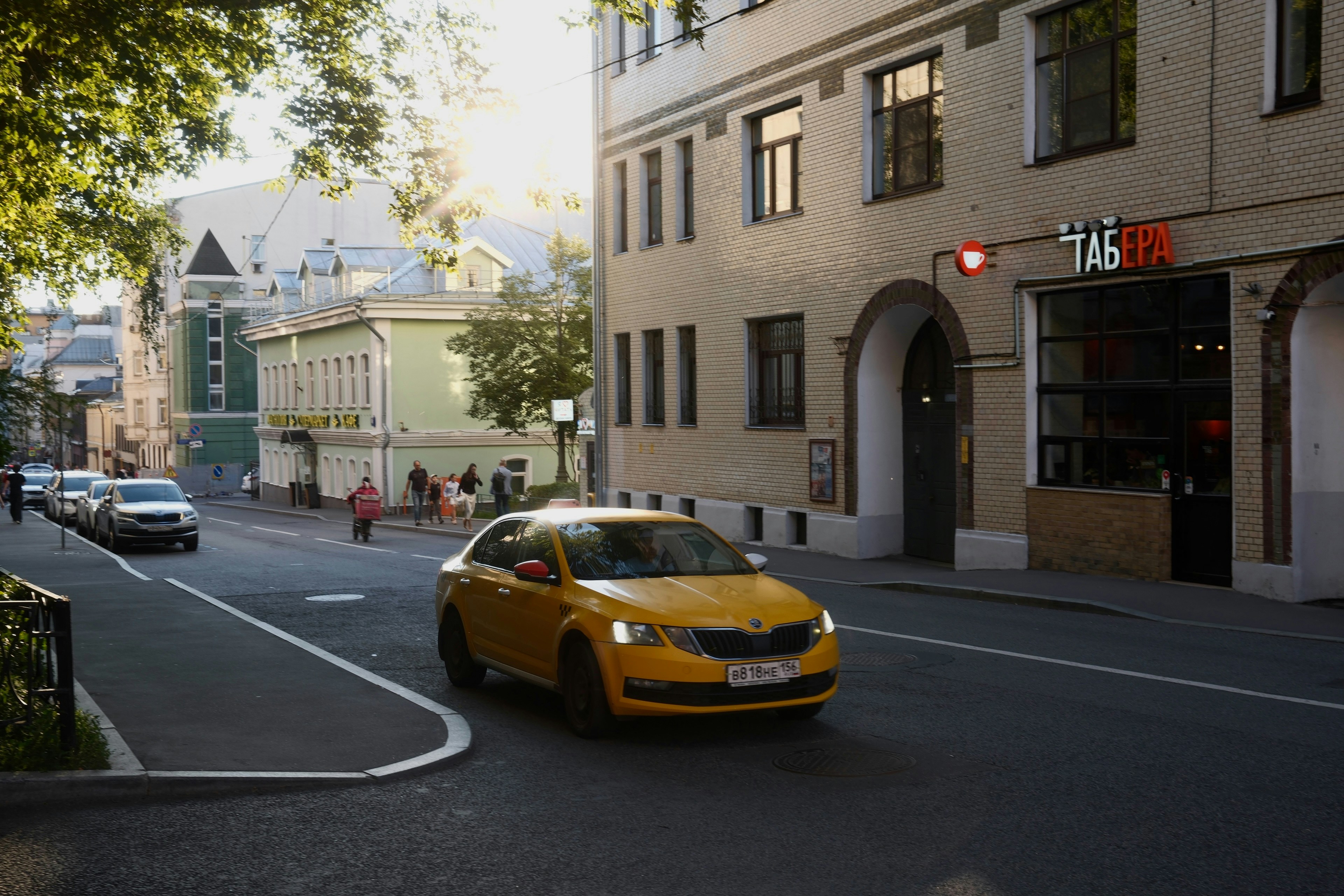 A yellow taxi driving on a city street past a brick building