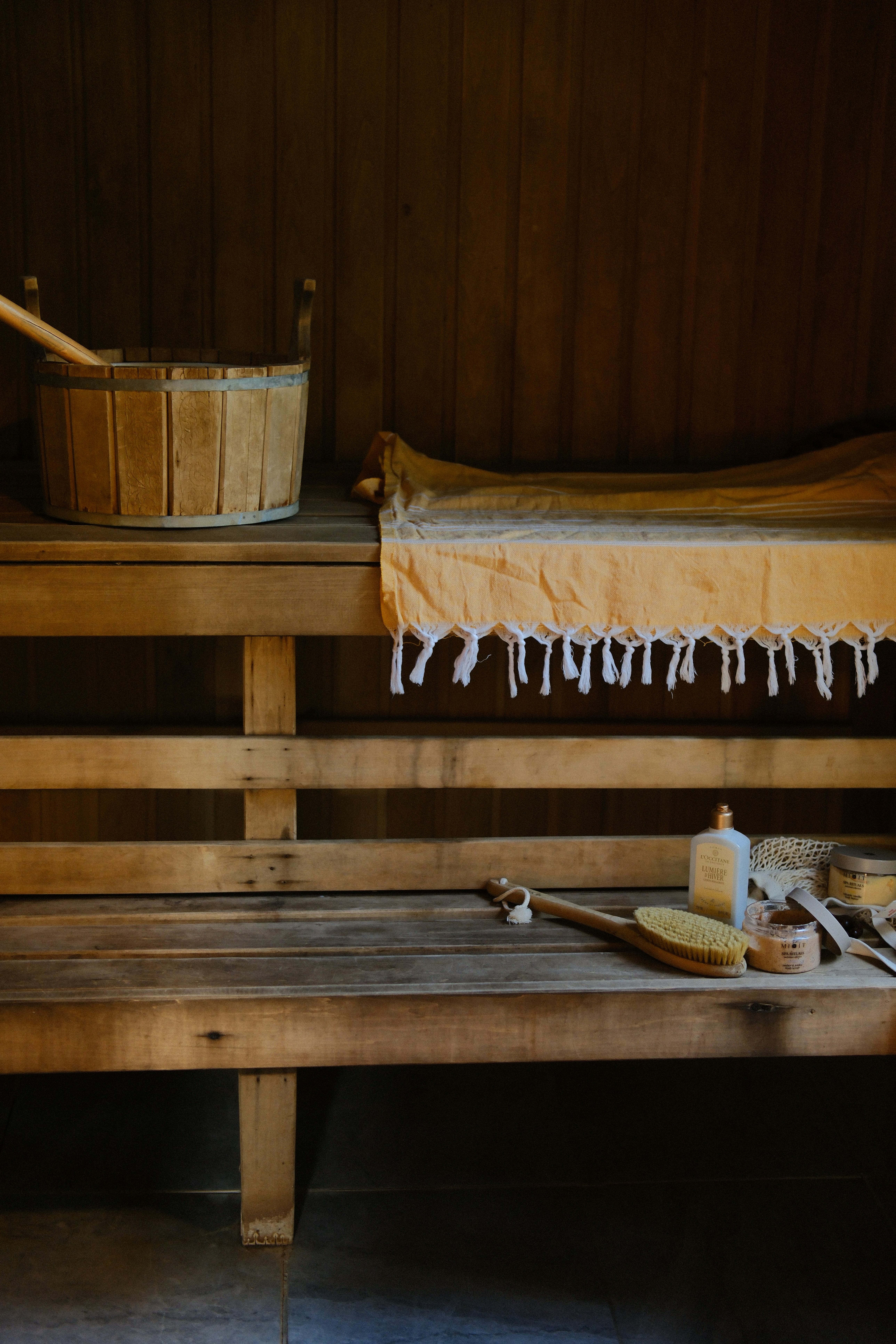A wooden sauna bench with a bucket, yellow towel, and bath accessories.