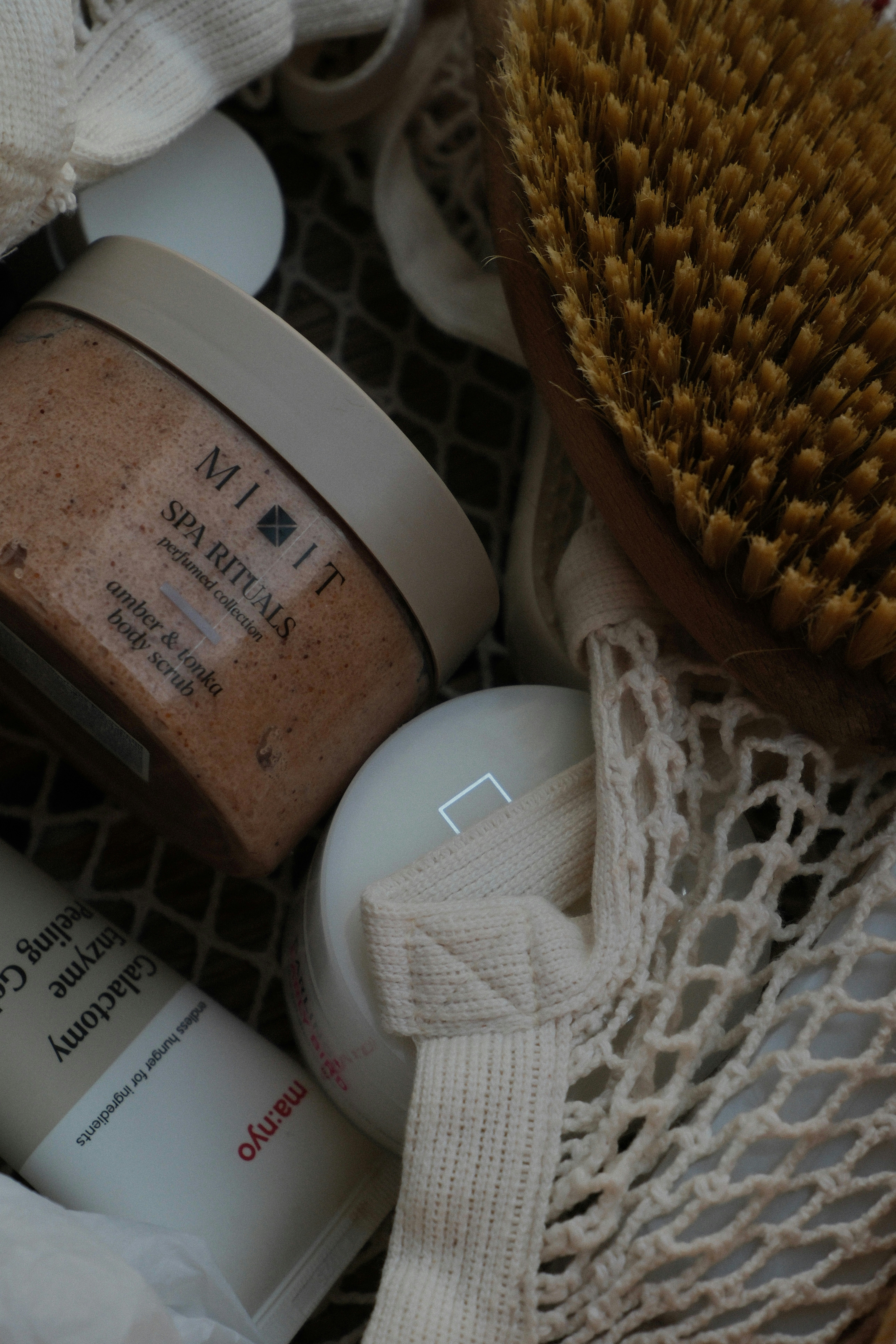 A body scrub jar, a wooden brush, and skincare products in a mesh bag