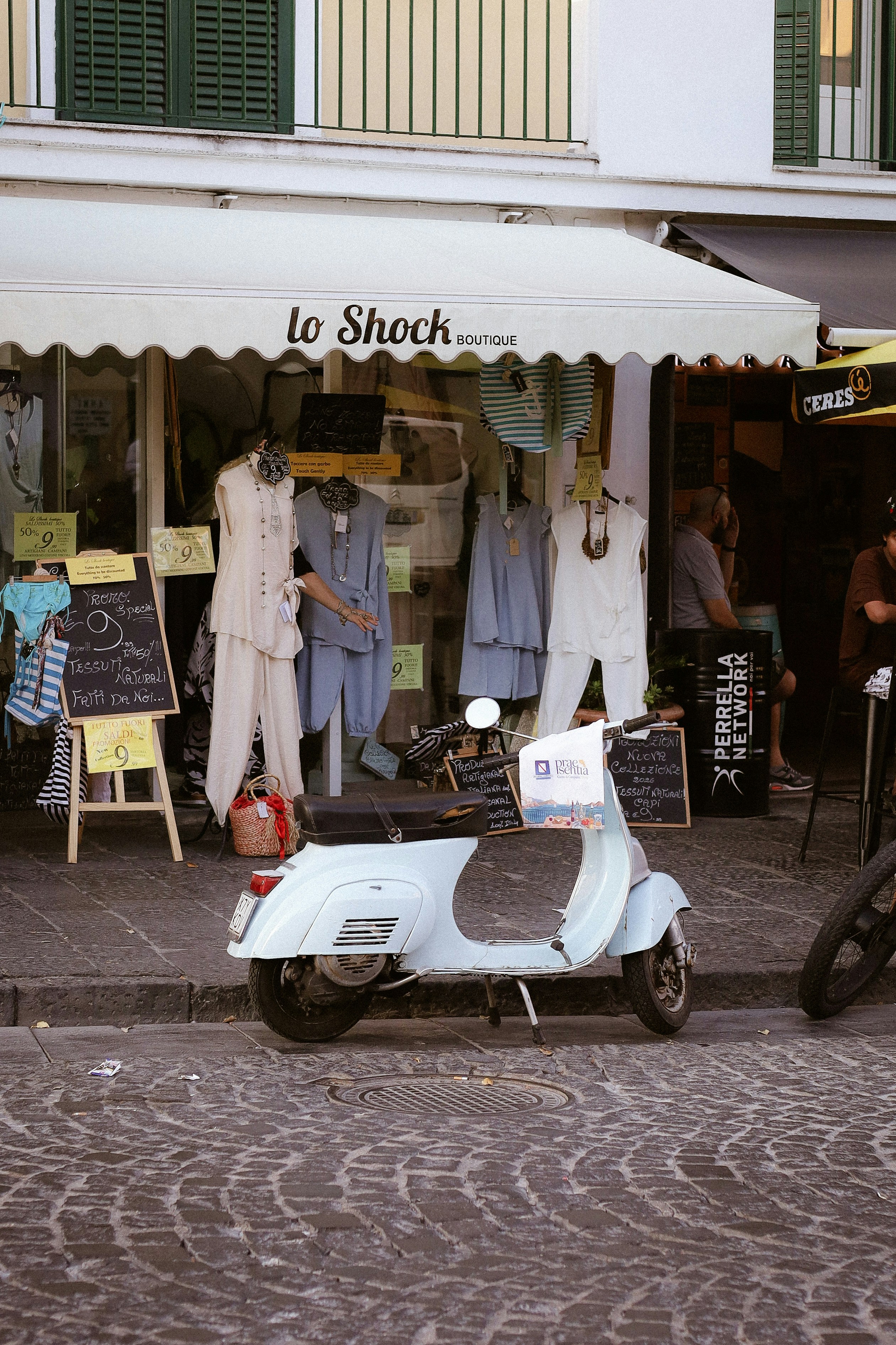 A light blue scooter parked on a cobblestone street in front of a boutique