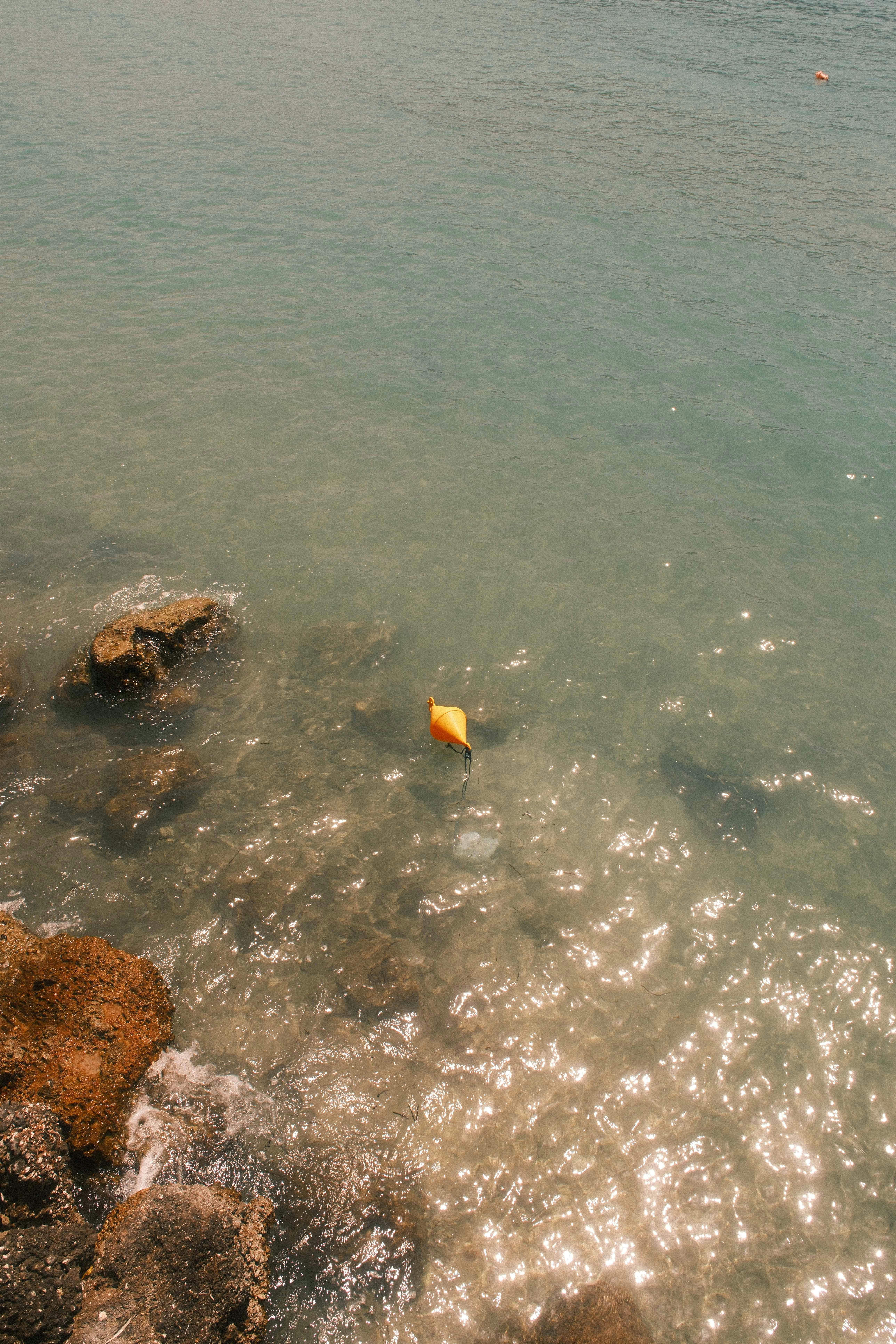 Pelampung kuning cerah mengapung di air jernih yang diterangi matahari dekat garis pantai berbatu.