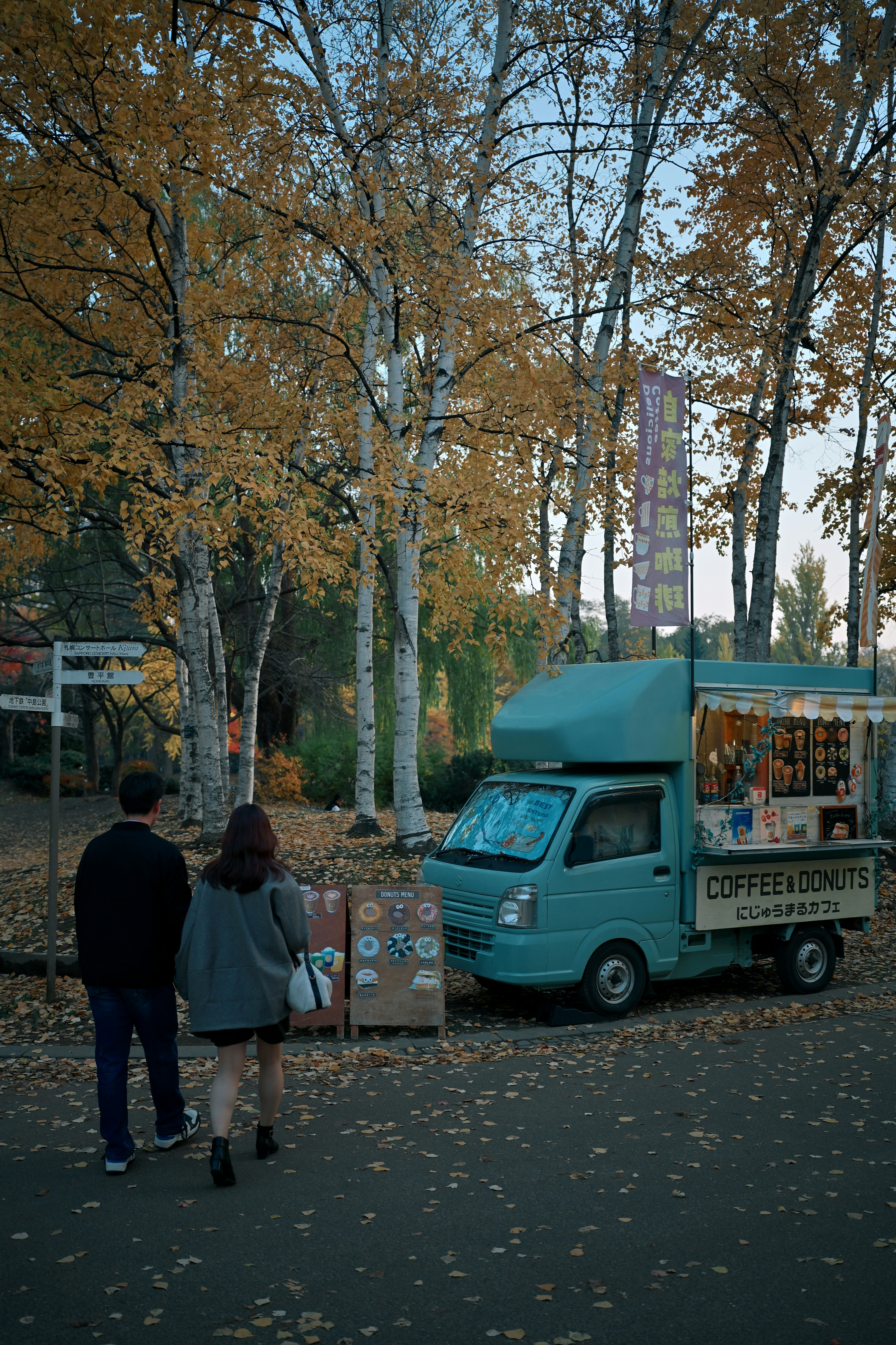 Two people walking toward a teal coffee and donut truck in a park.