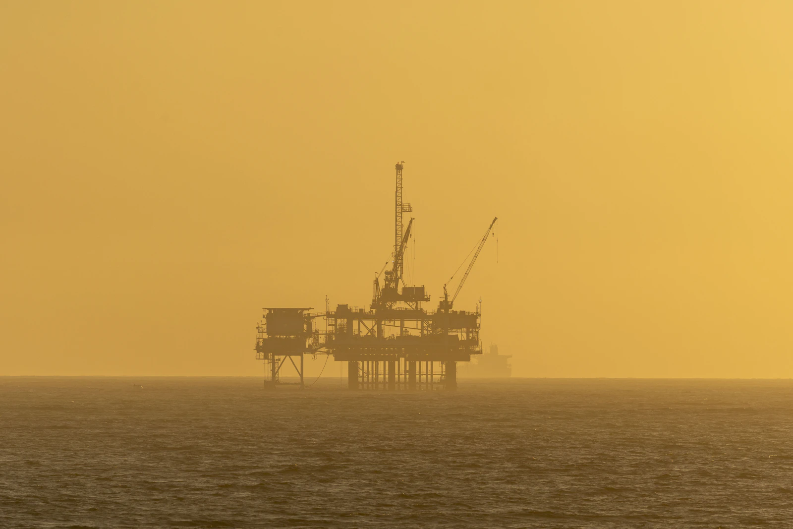An offshore oil rig in silhouette against a warm orange sky over the ocean