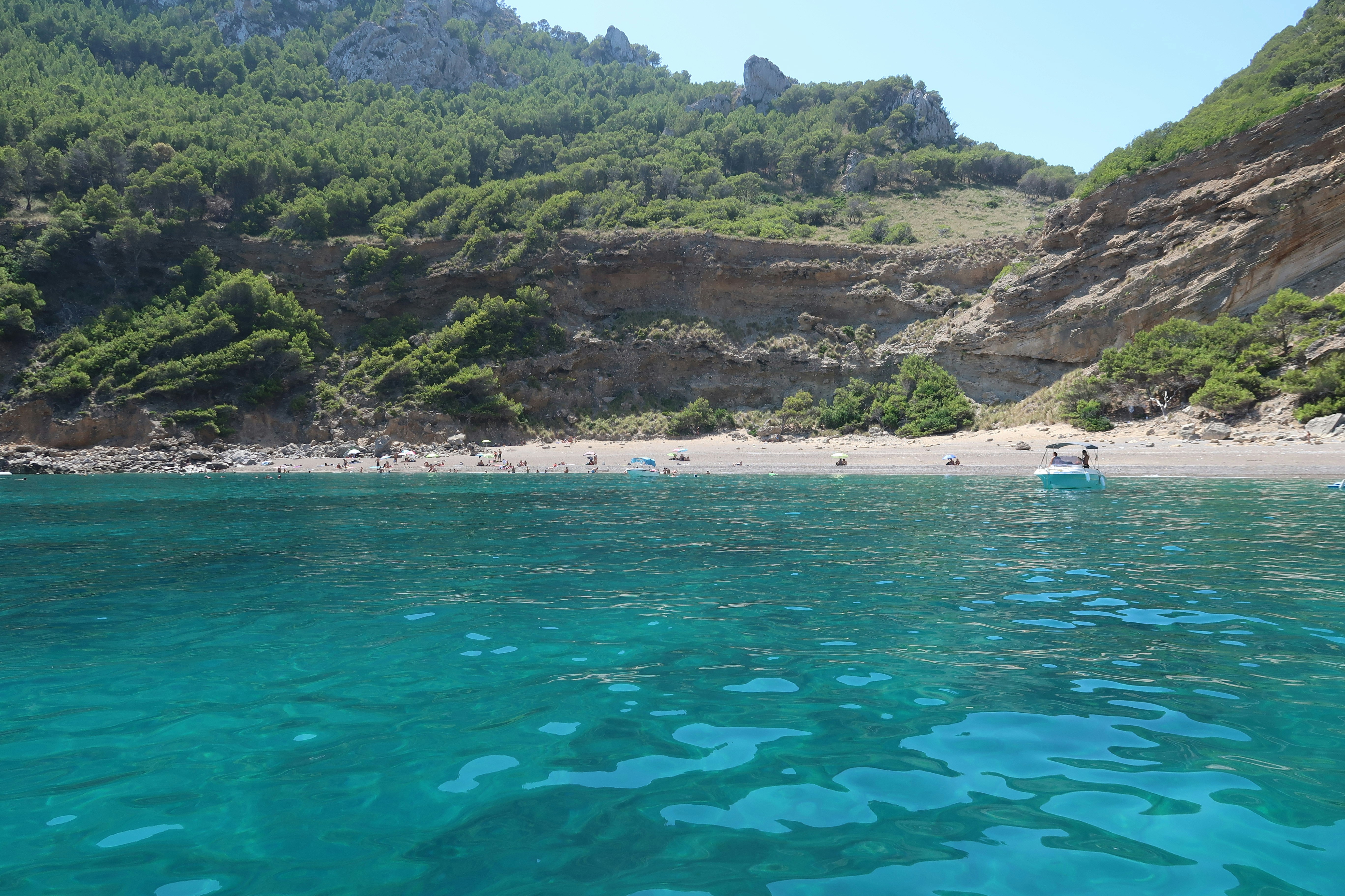 A turquoise bay with a pebble beach, rocky cliffs, and pine trees