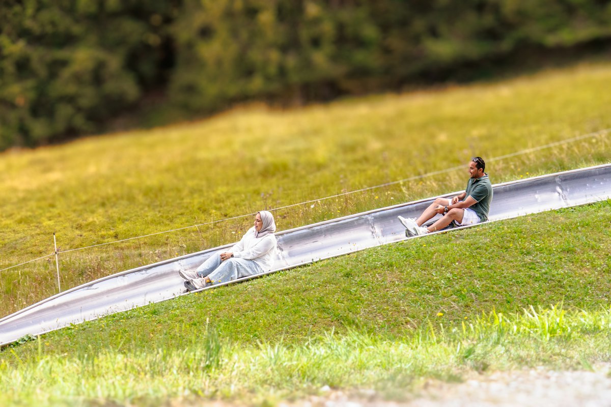 Two people in a single cart descending a metal rail that curves down an open grassy slope