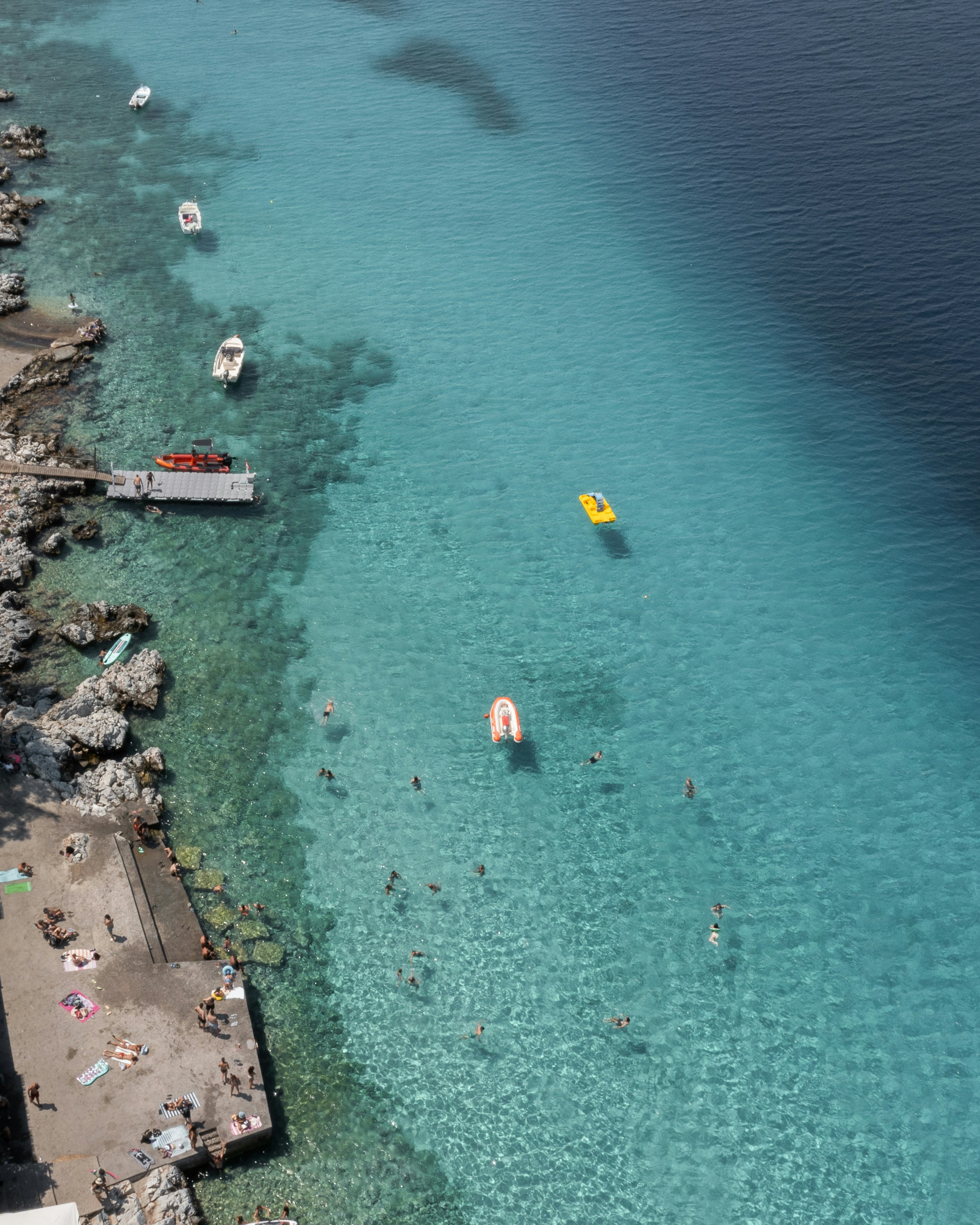 People swimming in clear turquoise water near a rocky shore with small boats