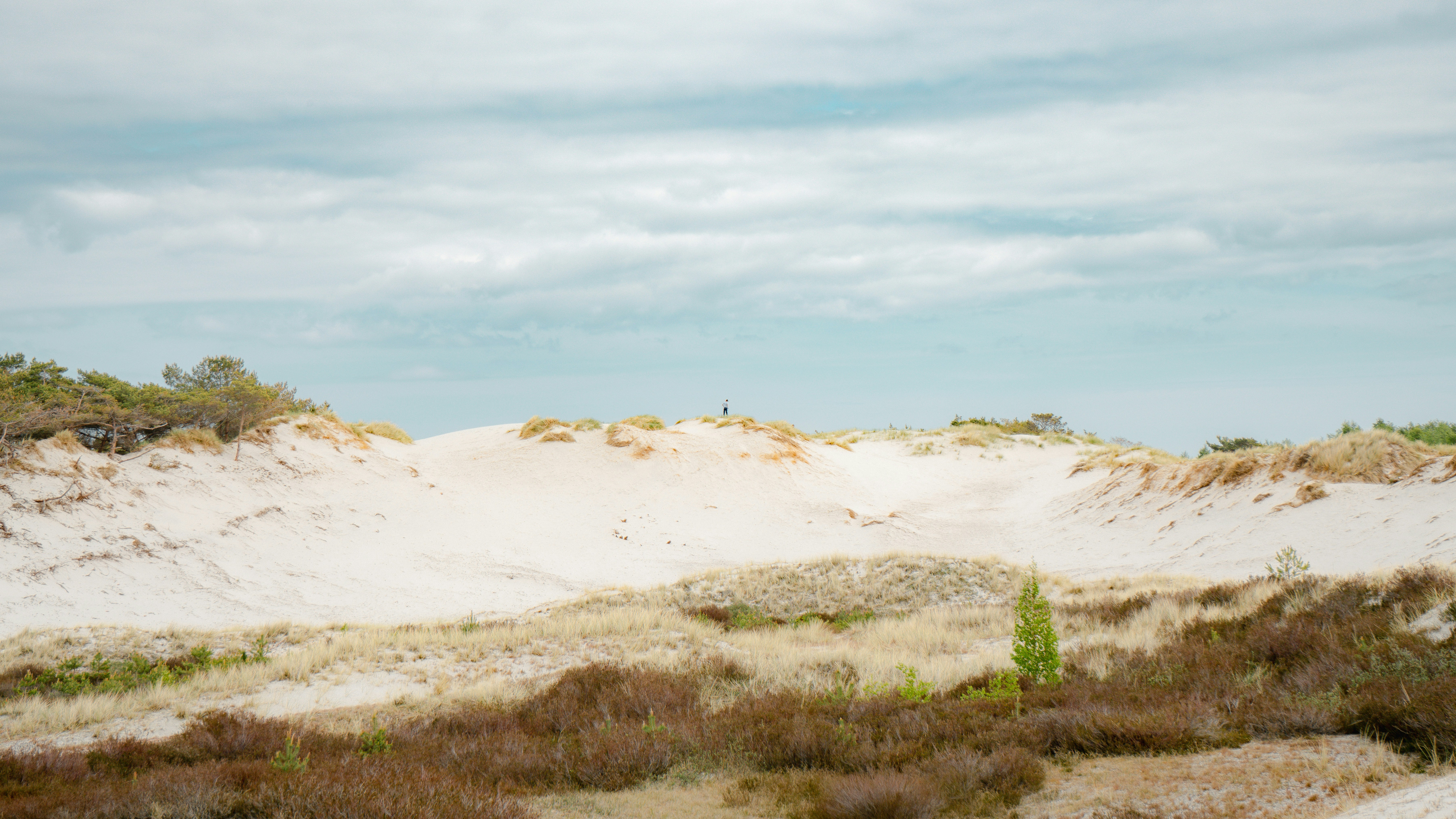 A person standing on a white sand dune with sparse grass under cloudy skies.