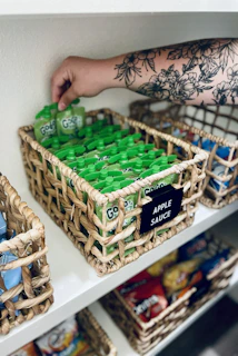 A person with a floral tattoo reaching for a green applesauce pouch in a basket
