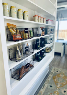 Black wire baskets on white shelves containing snacks, smoothie ingredients, and produce