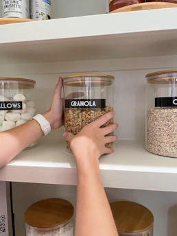 A person placing a glass jar labeled "GRANOLA" onto a white pantry shelf