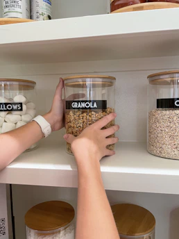 A person placing a glass jar labeled "GRANOLA" onto a white pantry shelf