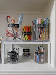 Clear plastic containers labeled with office supplies on a white shelf