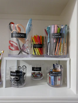 Clear plastic containers labeled with office supplies on a white shelf