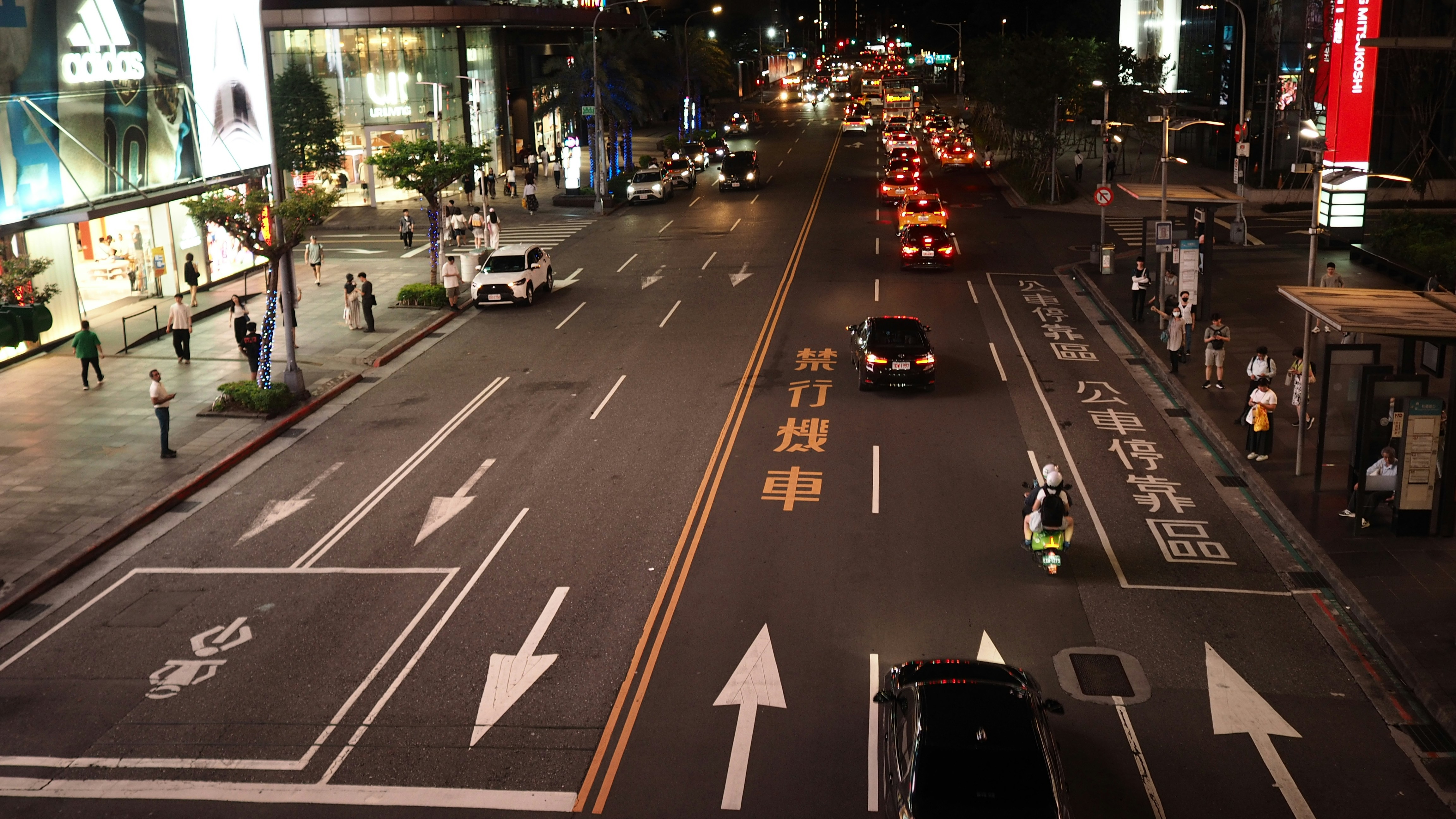 Cars and a scooter on a city street at night with illuminated storefronts