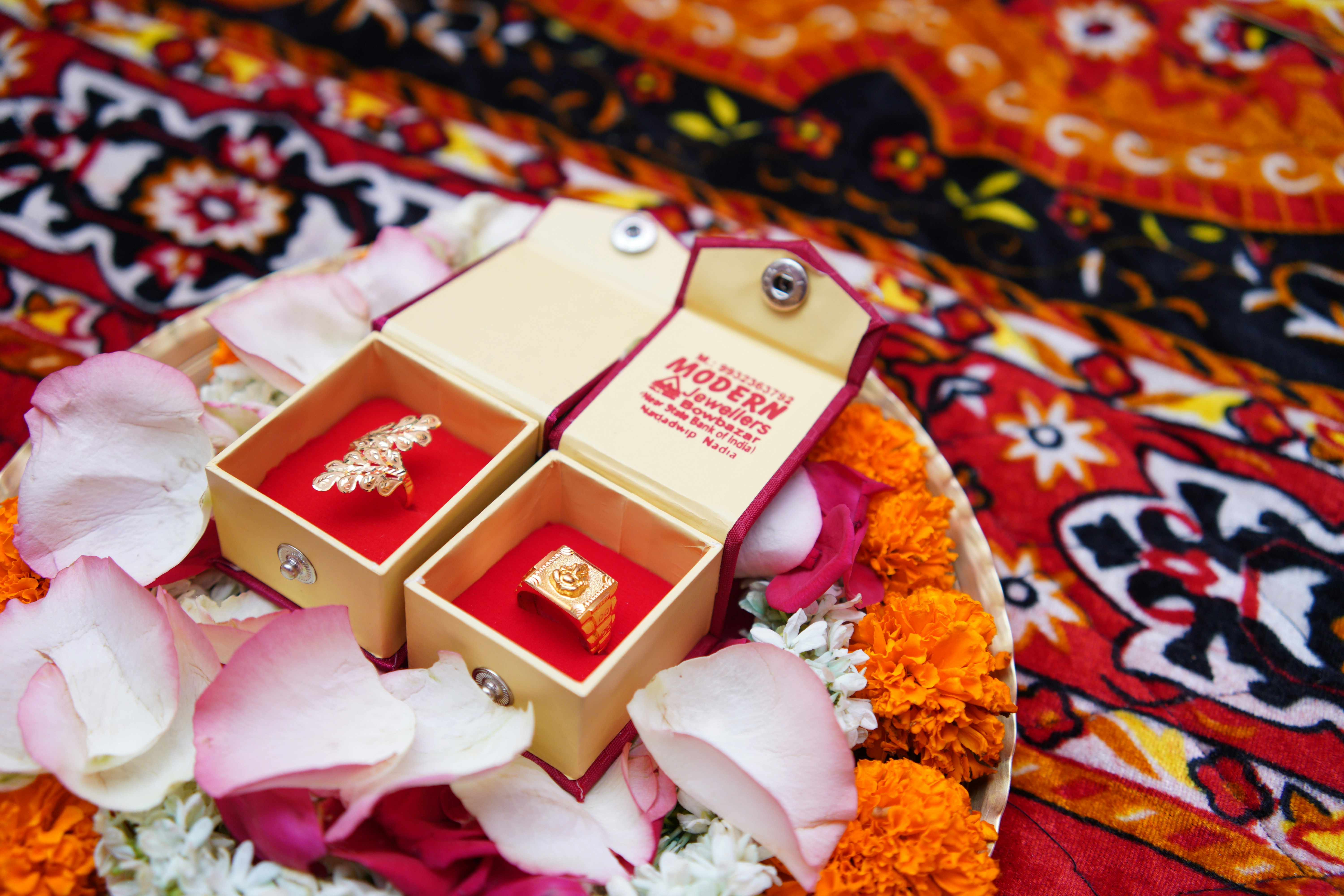 Two gold rings in open boxes surrounded by rose petals and marigold flowers