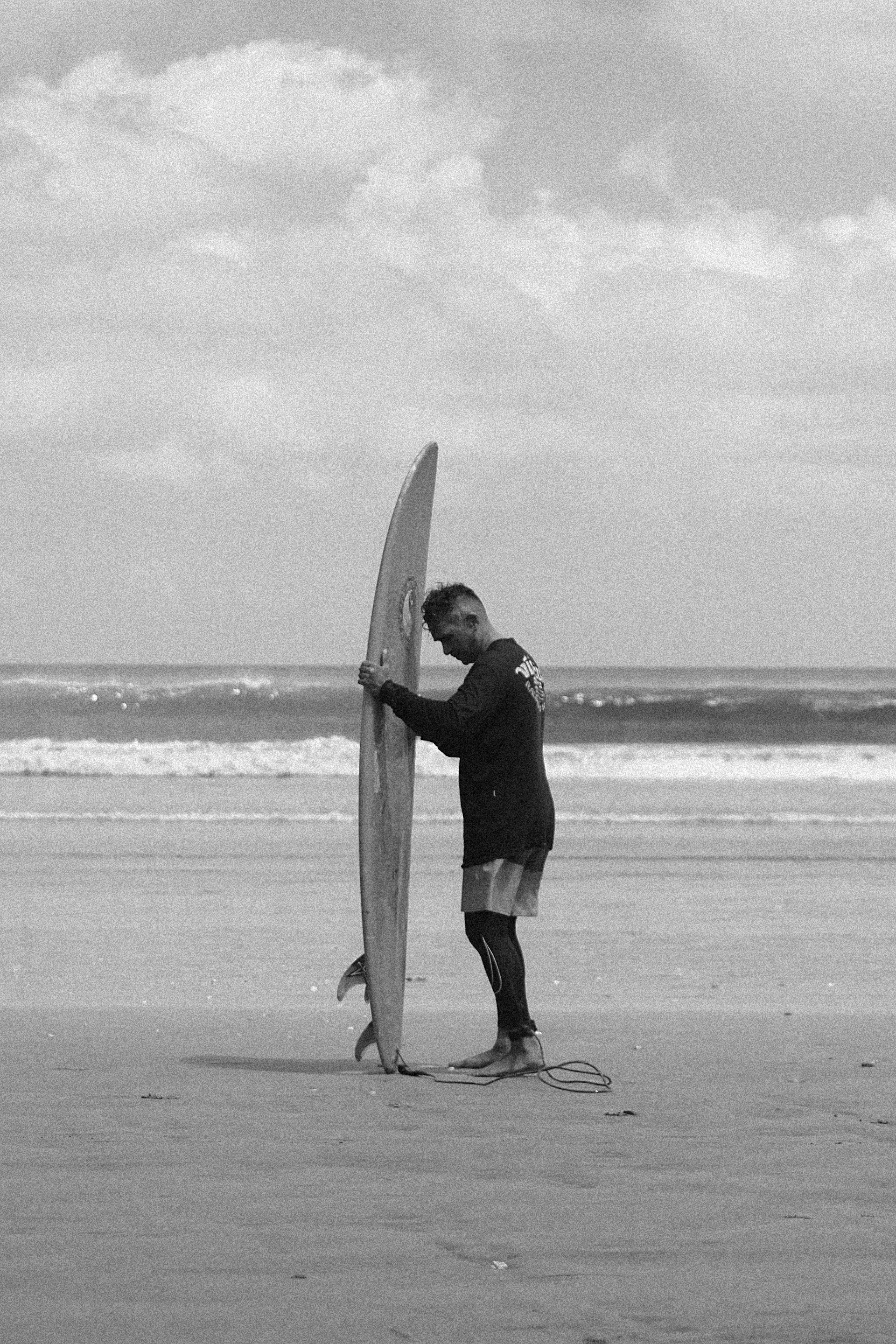 Eine Person in einem langärmligen Hemd hält ein Surfbrett an einem Sandstrand